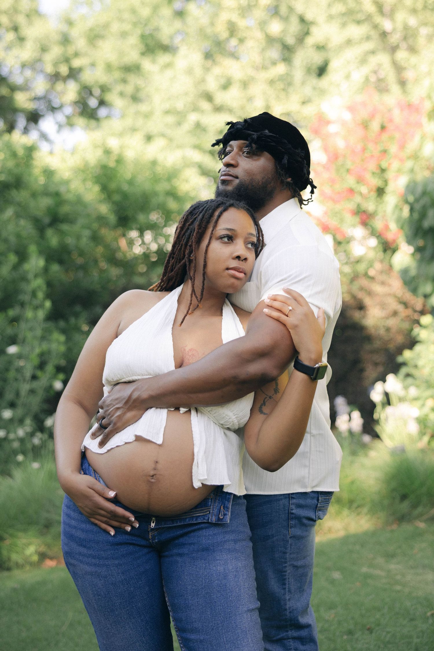Pregnant Black woman in white crop top and jeans embraced by partner outdoors in lush green garden setting.