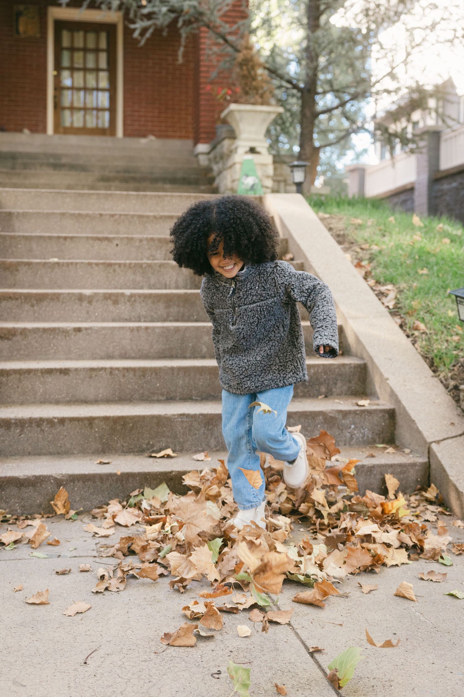 A small child in a sweater and jeans plays in a pile of fallen autumn leaves on concrete steps.