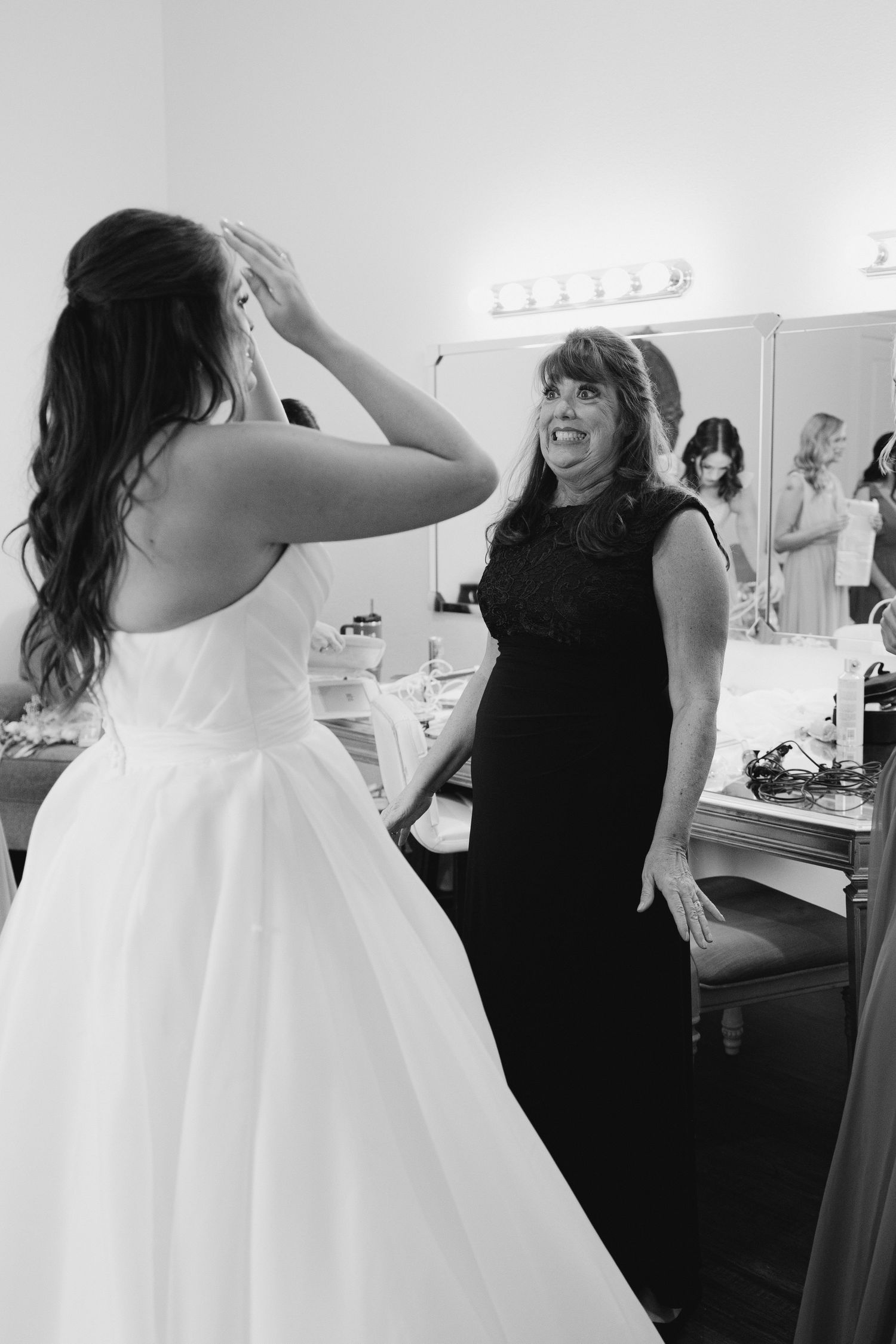 A bride in a white wedding dress gets help with final adjustments in a dressing room.