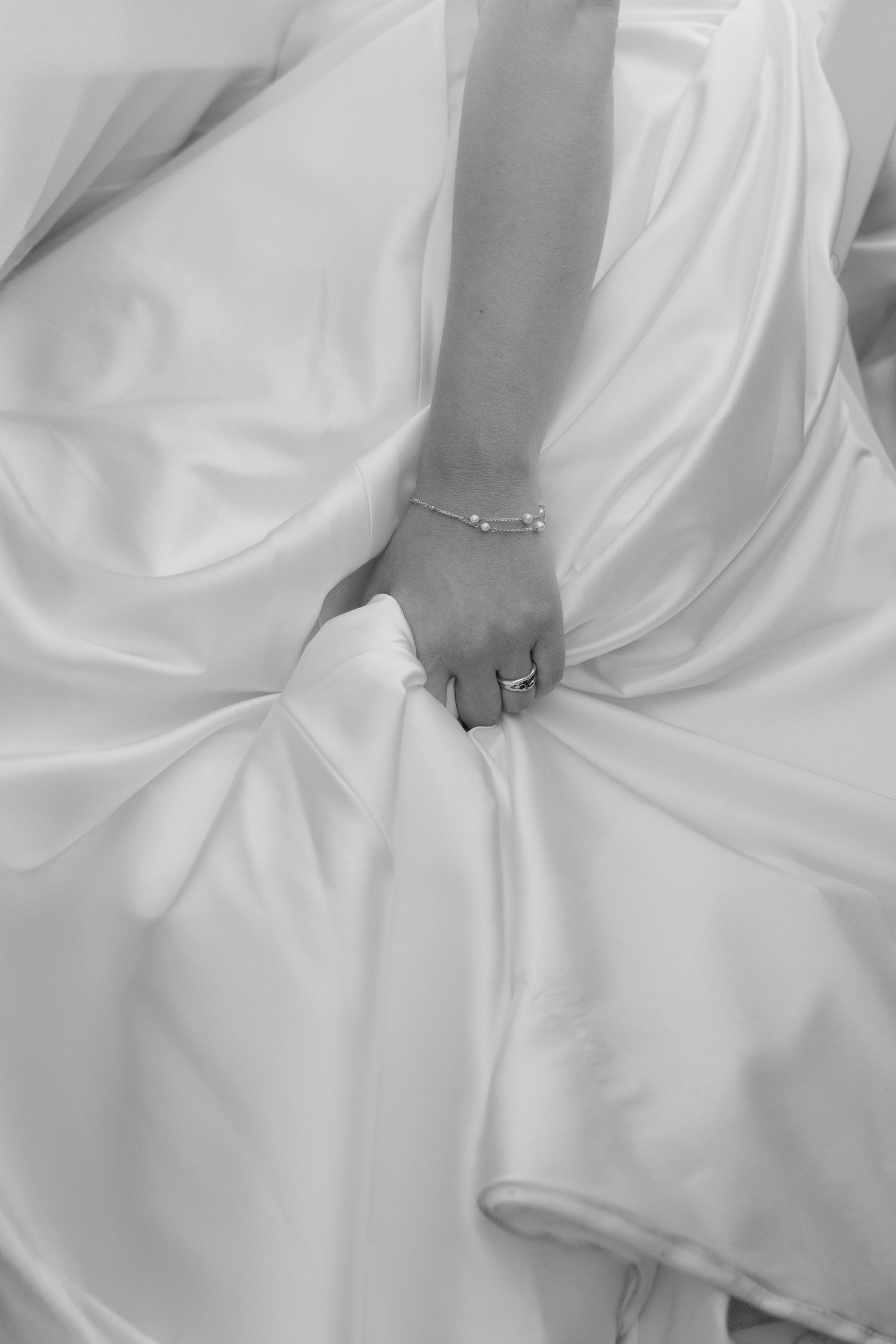 Black and white close-up of bride's hand holding white wedding gown, wearing diamond ring and bracelet.