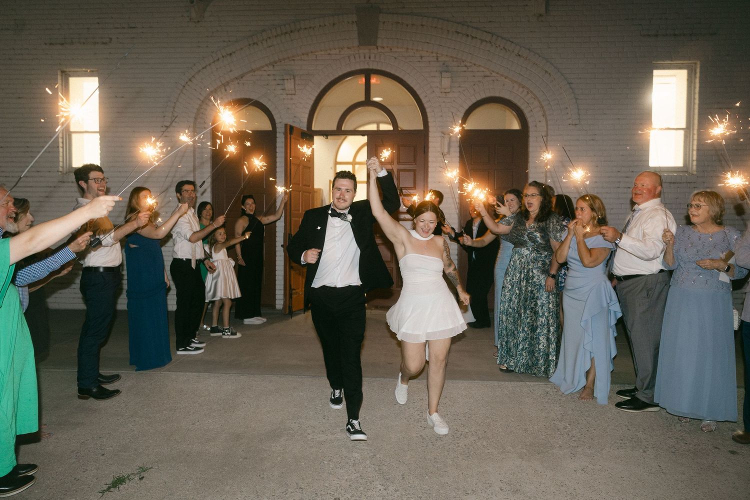 Bride and groom exit church through sparkler send-off with cheering guests lining the path at night.