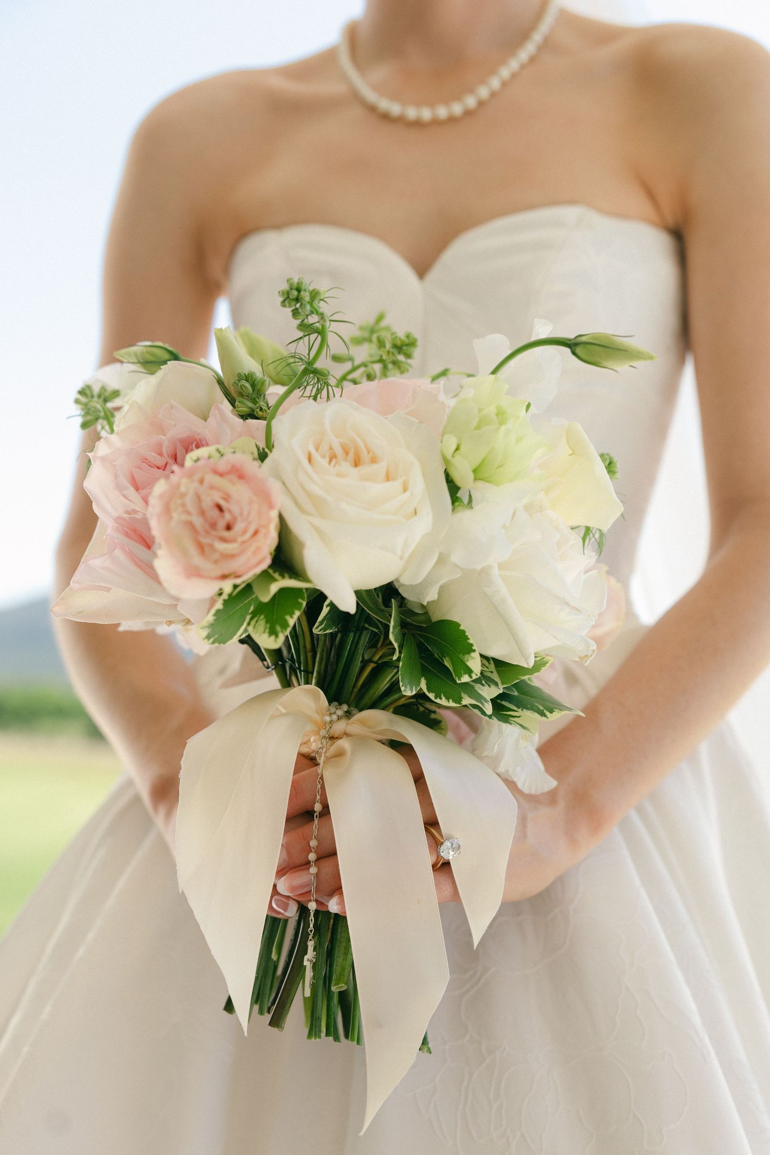 Bride holding elegant bouquet of pink and white roses with ribbon at outdoor wedding.