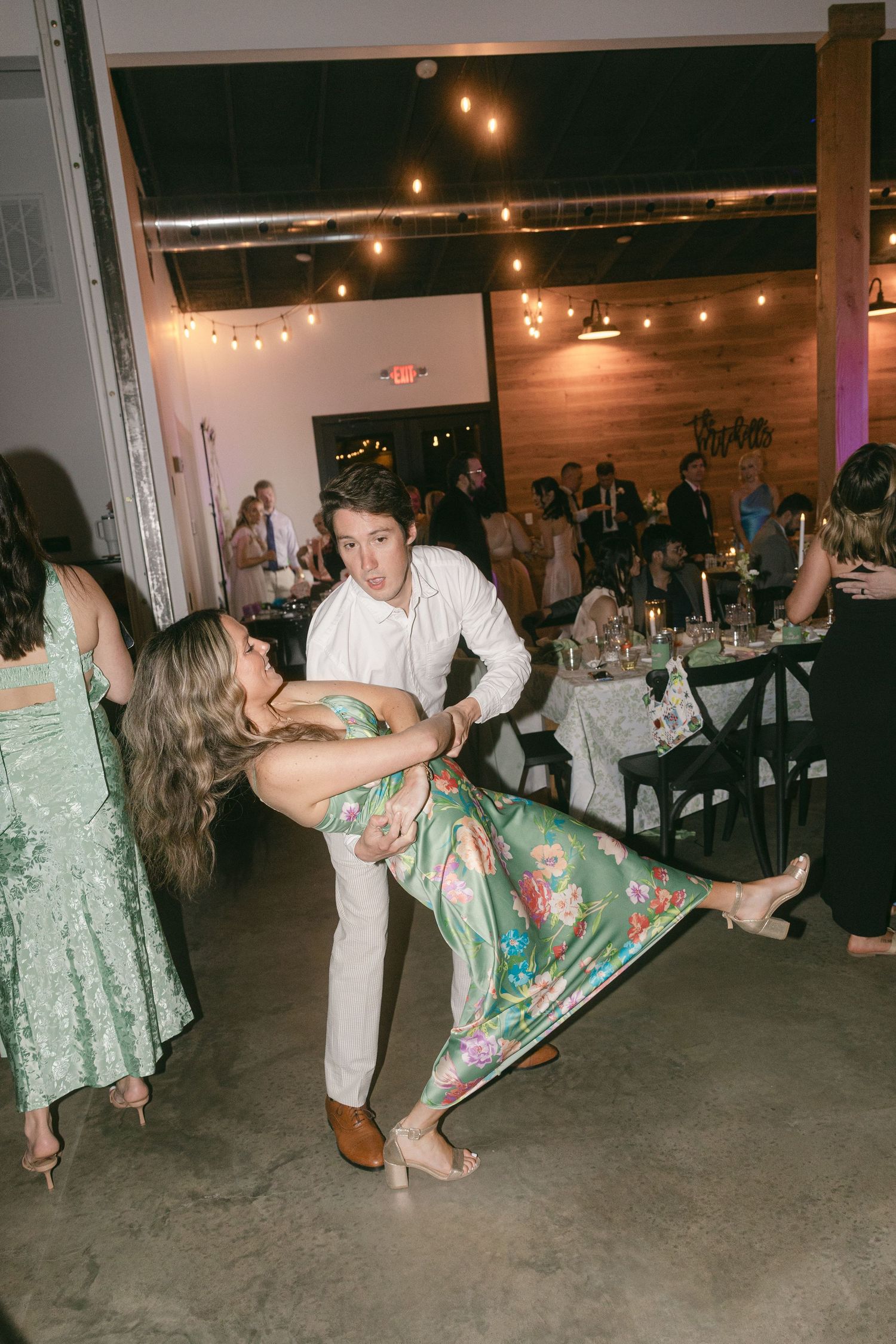 A couple dancing at a wedding reception, with the man dipping the woman in a floral green dress under string lights.