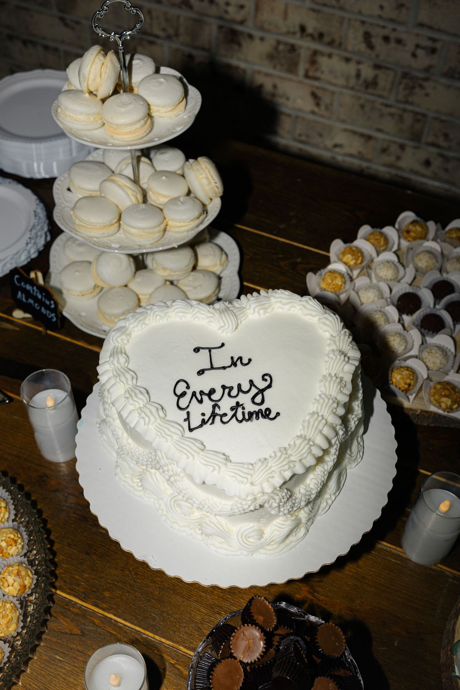 White wedding cake with romantic text inscription sits on display next to macarons and other desserts on wooden table.