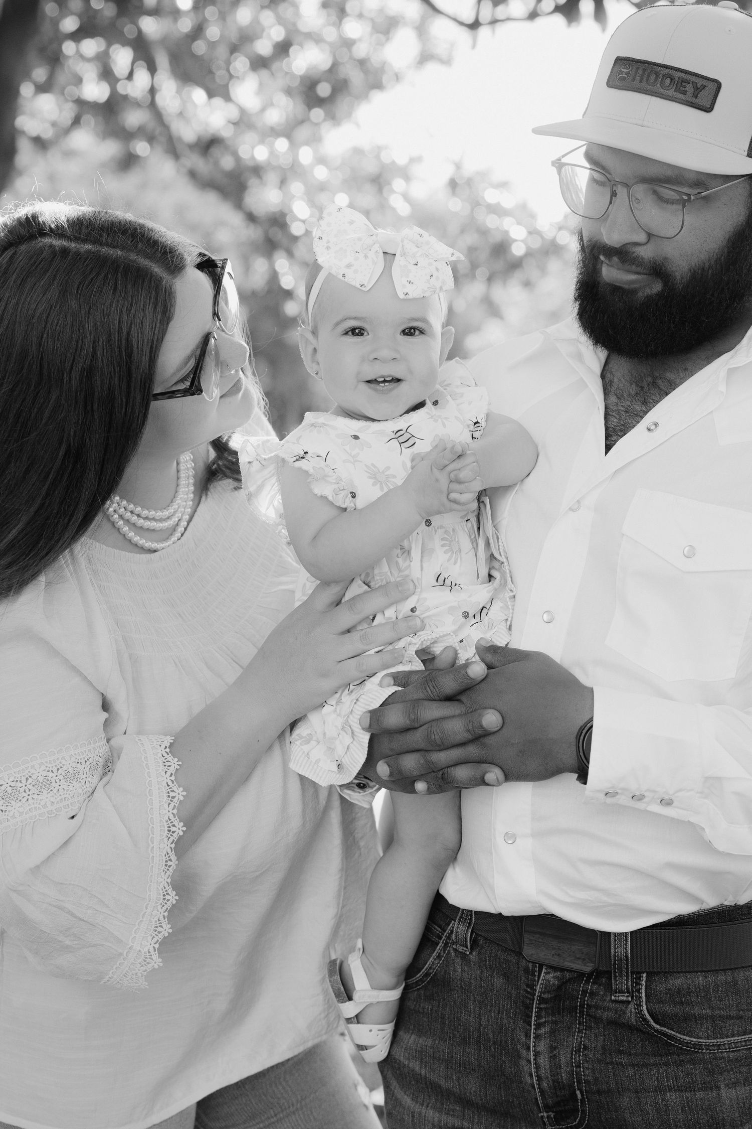 Black and white outdoor family portrait showing a couple with a small child wearing a white dress against a soft bokeh background.