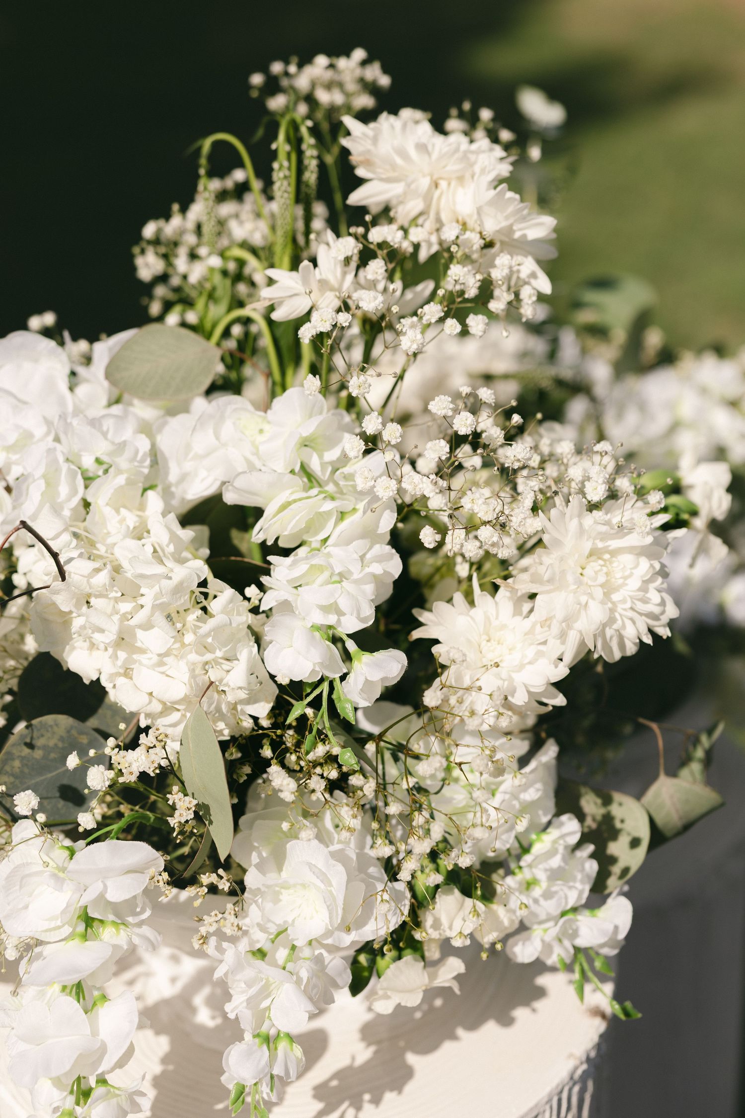 Elegant white wedding bouquet featuring dahlias, gypsophila, and orchids with lush greenery in natural sunlight.