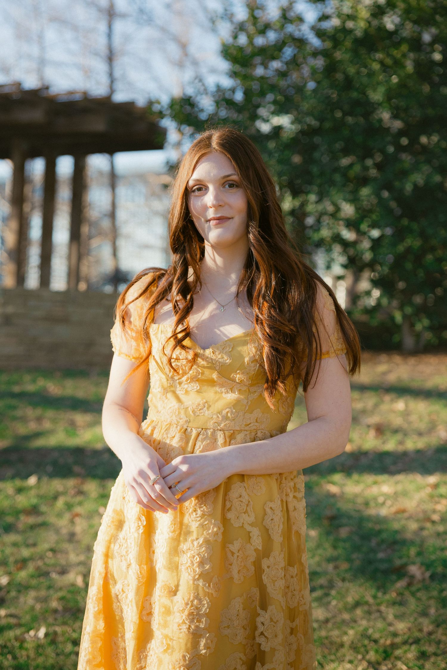 Woman in yellow floral dress poses outdoors in natural setting with trees and greenery in background.