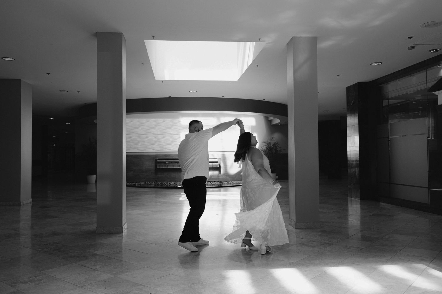 Black and white photo of a couple dancing together in an elegant indoor venue with marble floors and columns.