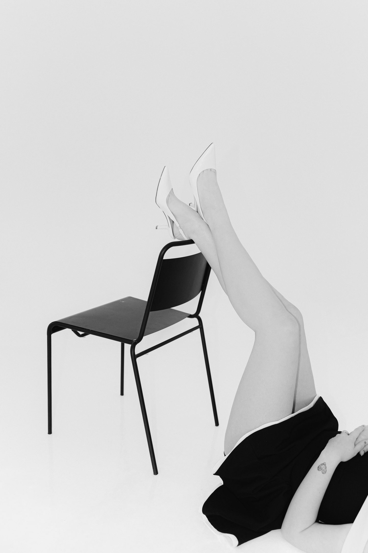 Black and white photo of legs raised against a black chair wearing white heels on white background.