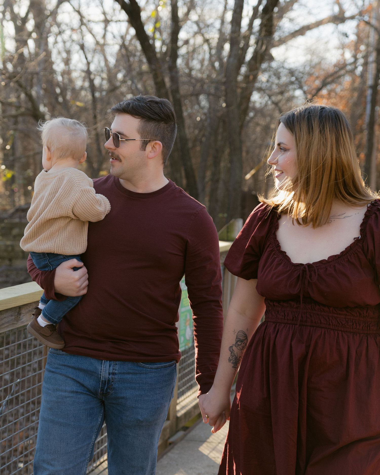 Family wearing coordinated burgundy outfits shares joyful moments along a wooden fence at sunset.