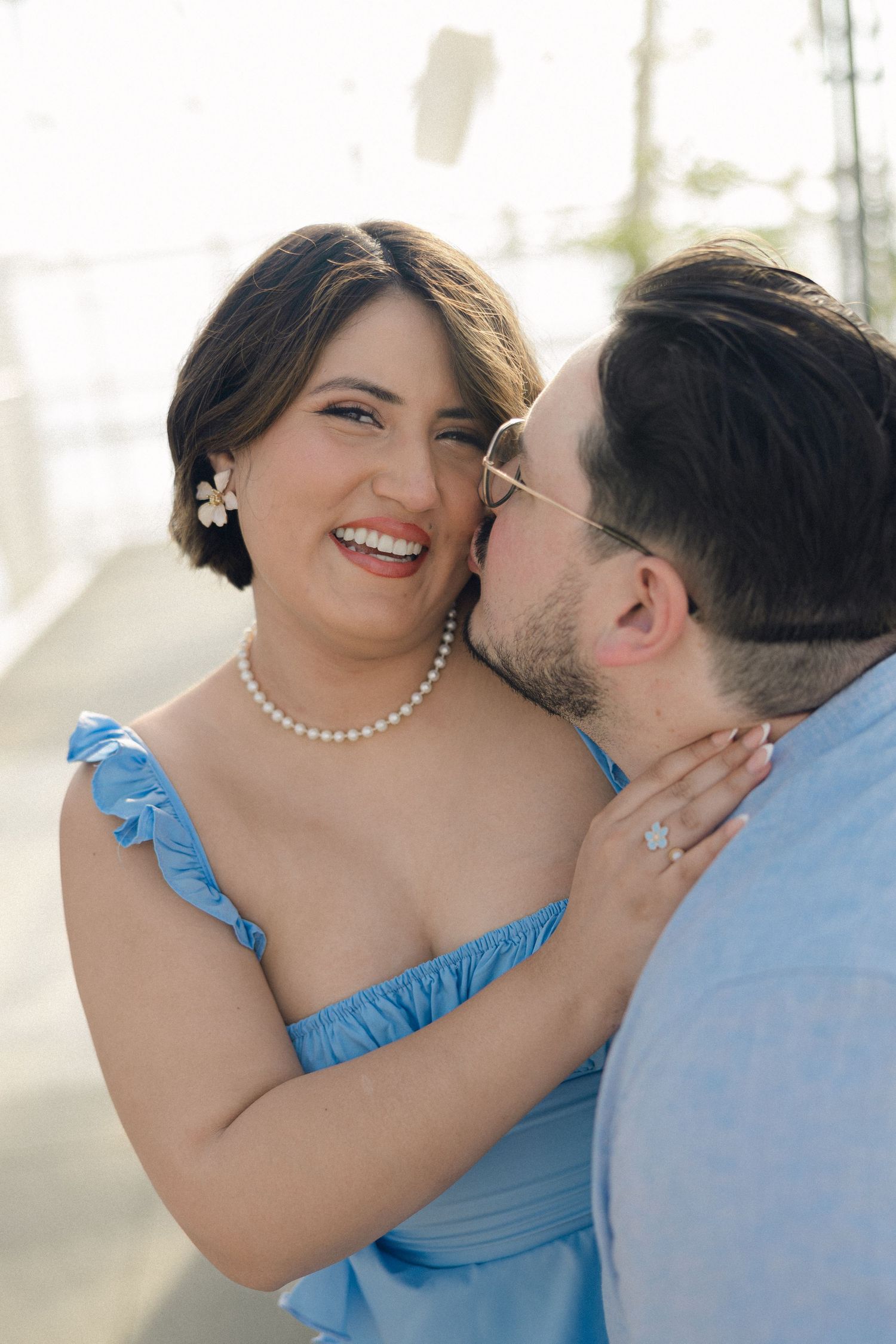 A smiling woman in a blue dress with pearl necklace embraces a man kissing her cheek during an engagement photo session.