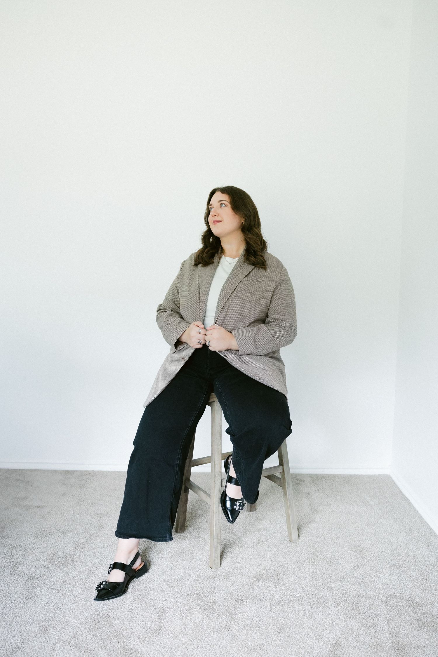 A person in a gray blazer and dark pants sits on a light colored chair in front of a white background striking various poses.