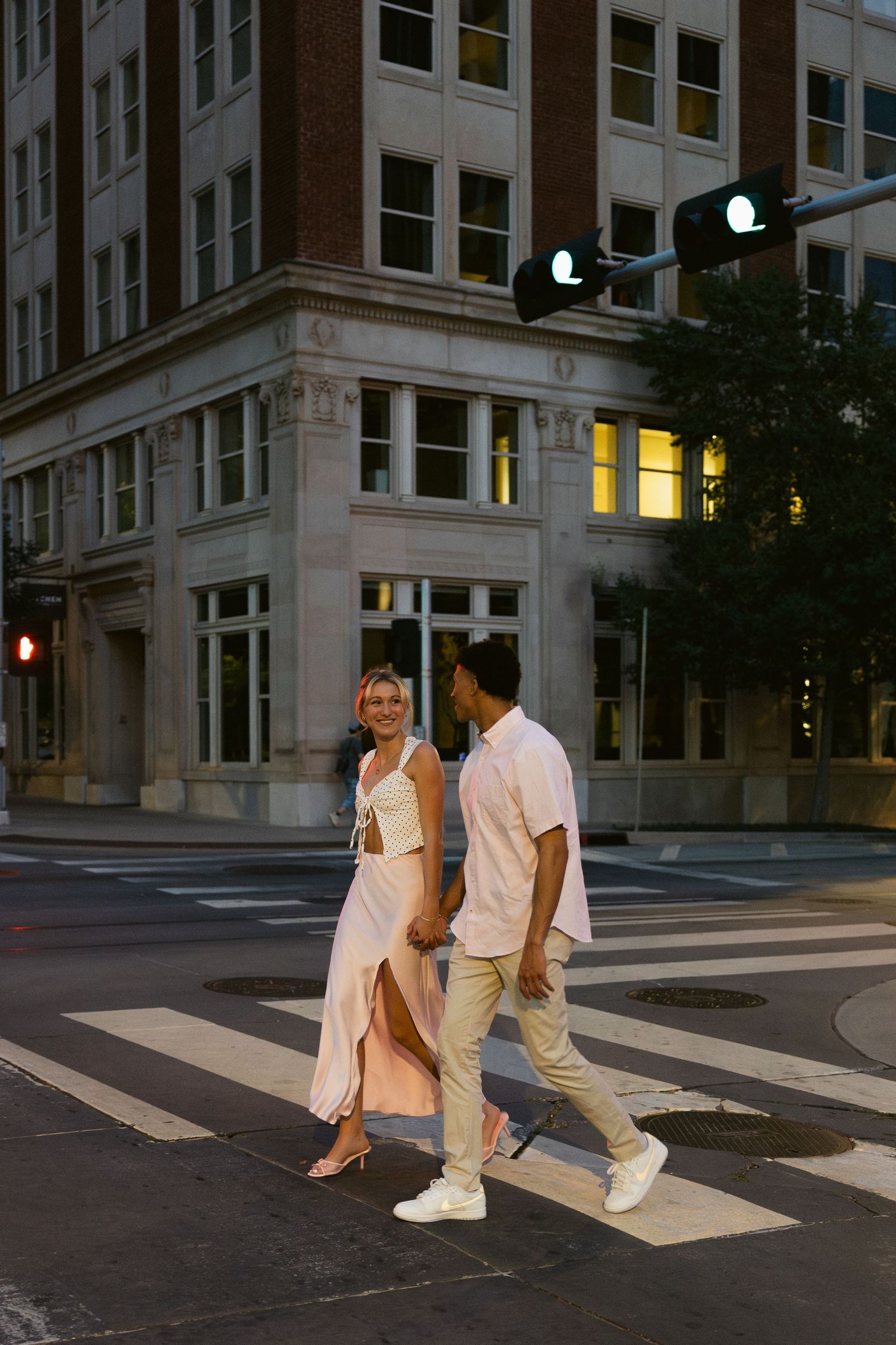 A couple in white outfits walks together across a crosswalk at dusk in an urban setting.