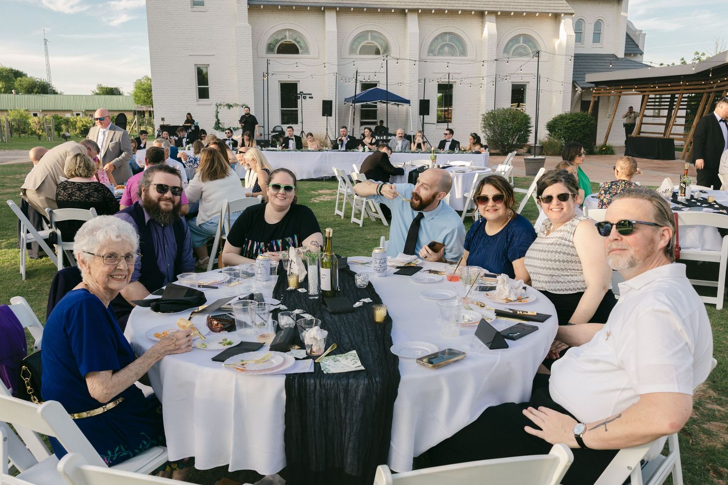 Group of guests smiling at round table during outdoor evening reception with white building backdrop.