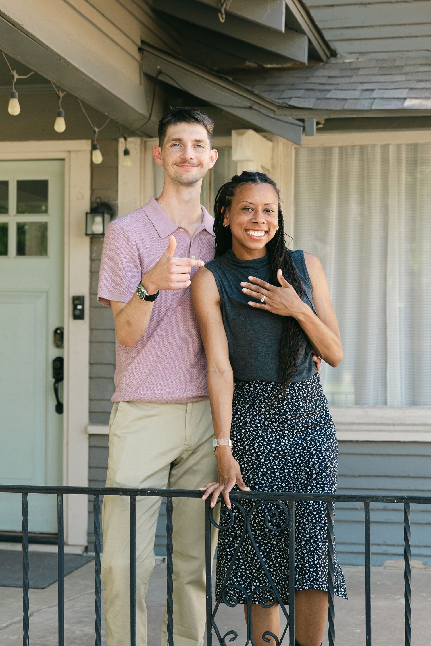 A happy interracial couple poses on a home porch, man giving thumbs up in pink polo, woman in black top and floral skirt.