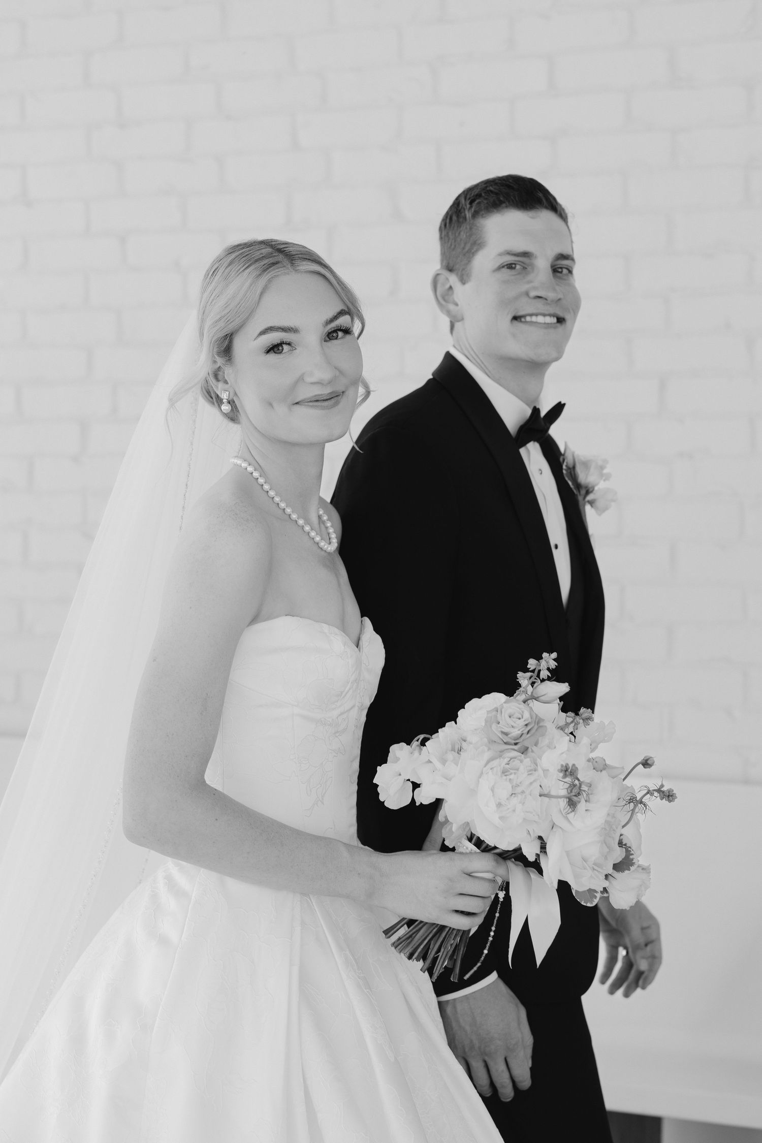 Black and white portrait of smiling bride holding bouquet with groom in tuxedo against white brick wall.