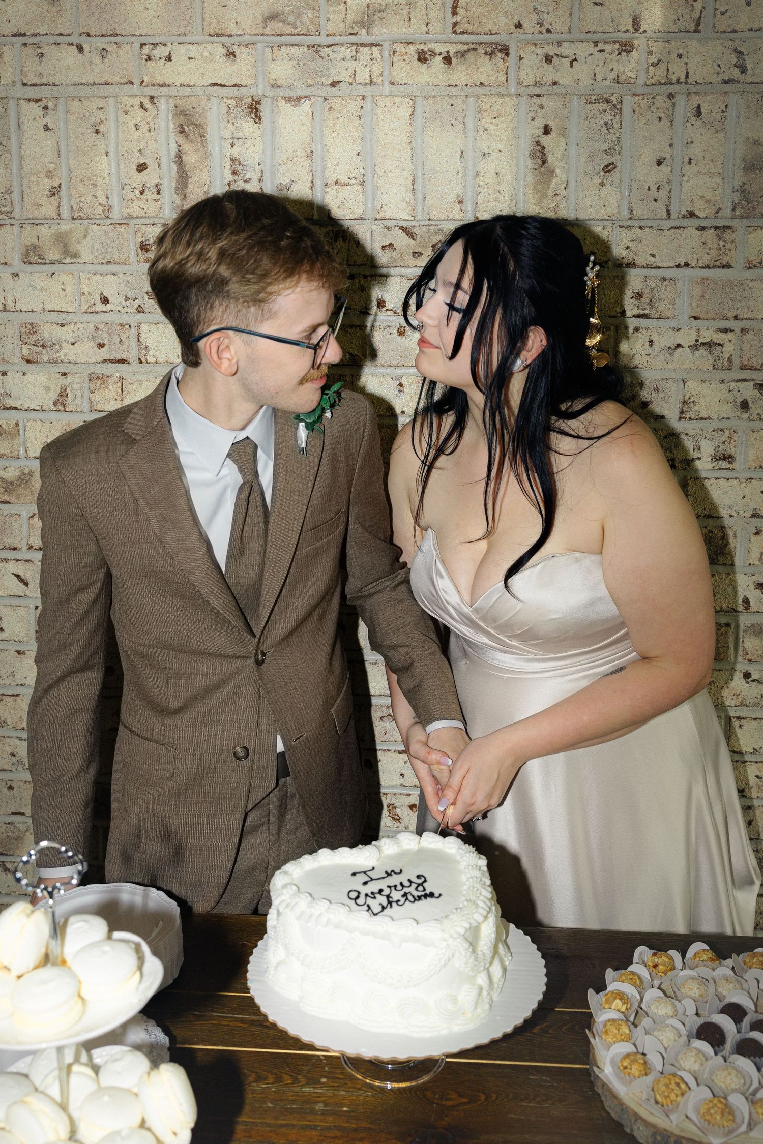 Newlyweds playfully share wedding cake at reception with white brick wall backdrop.