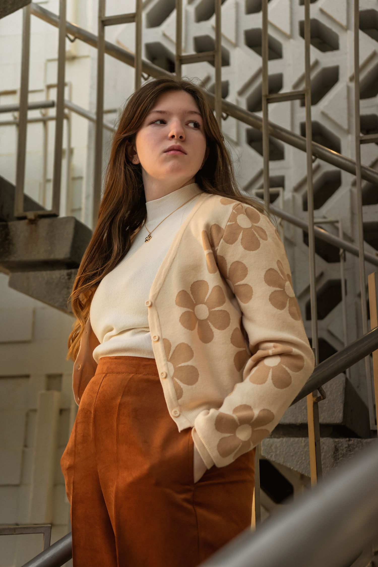 Retro-style fashion photo featuring cream floral sweater and rust-colored pants against industrial stairs.