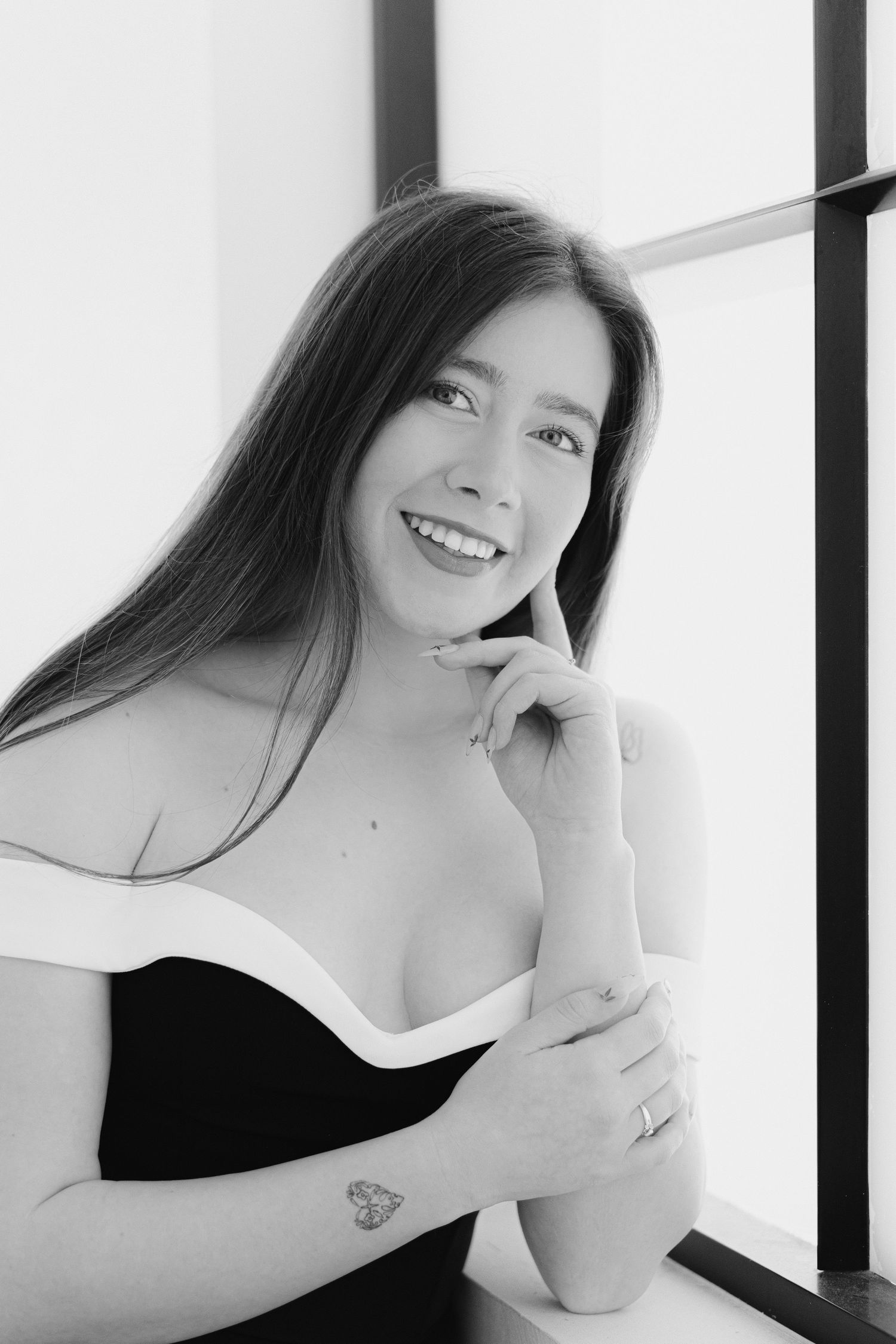 Black and white portrait of a smiling young woman with long hair, wearing an off-shoulder top, posing near a window.