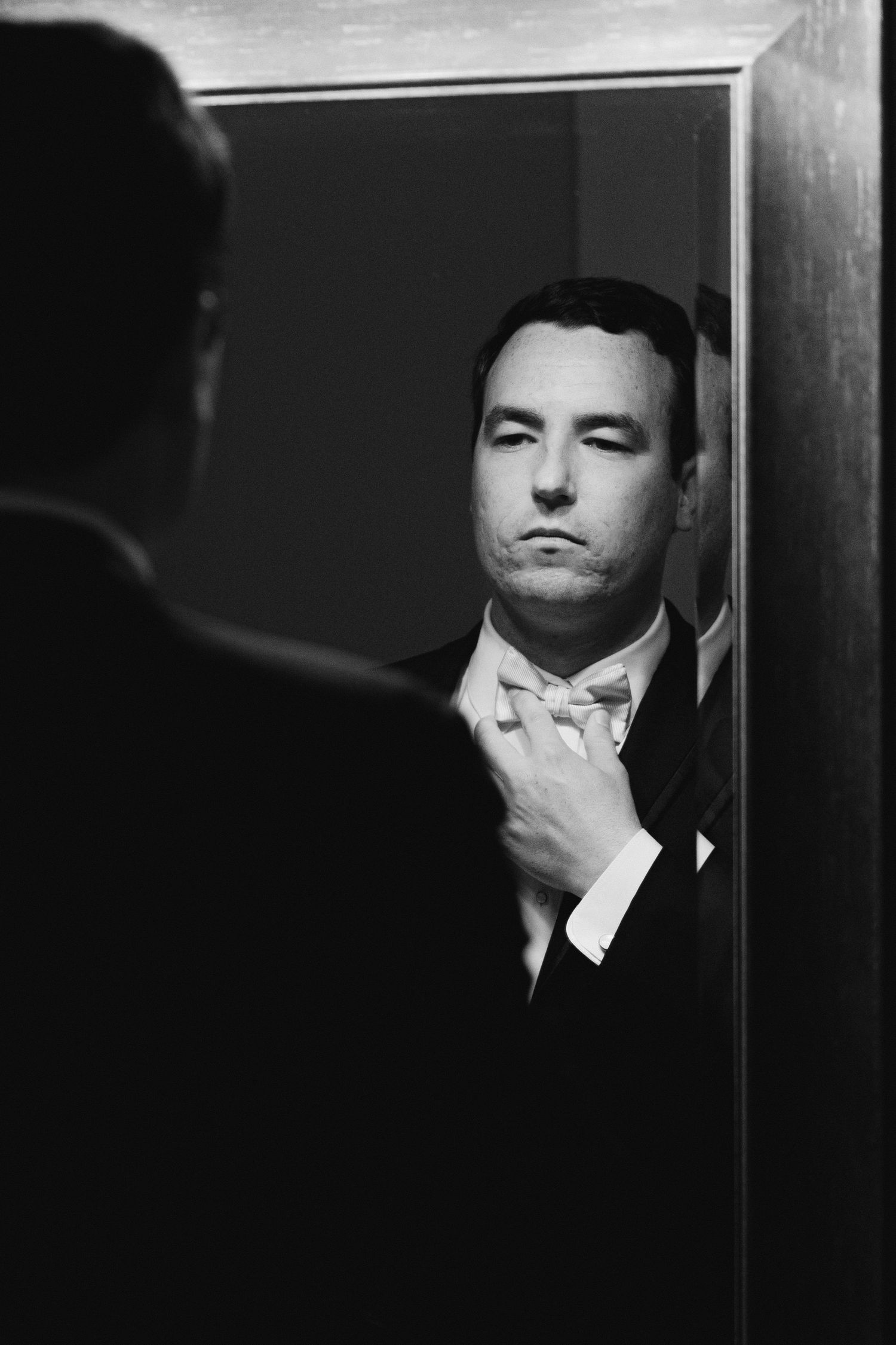Black and white reflection of a man adjusting his bow tie in a mirror while wearing a formal suit.