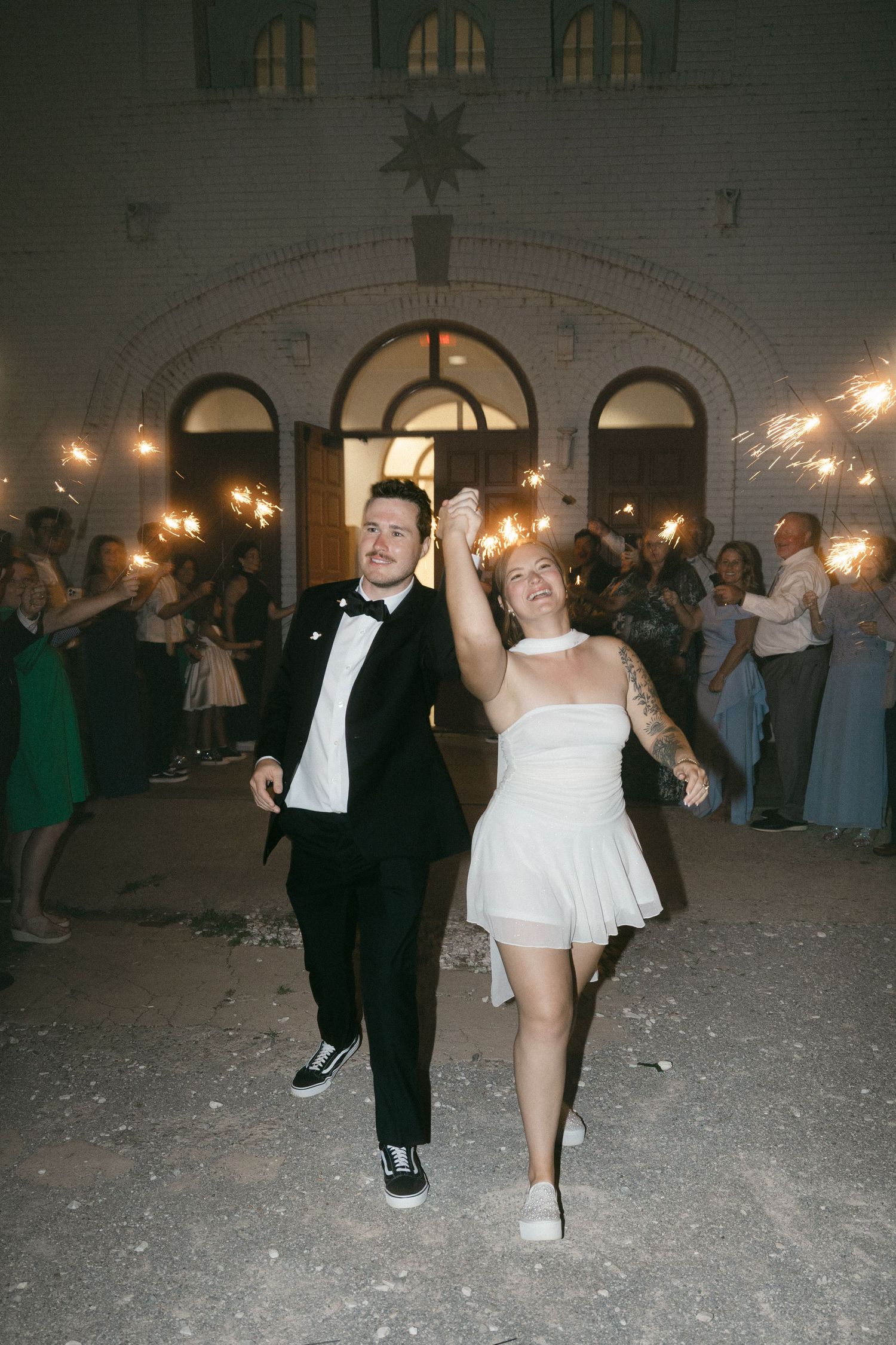 Bride and groom exit wedding venue through sparkler tunnel, bride in short white dress, groom in black tuxedo.
