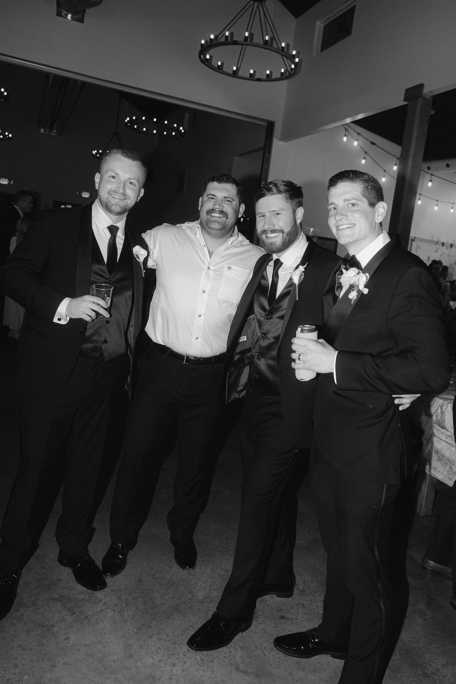 Four men in black suits smile together at wedding reception in black and white photo.