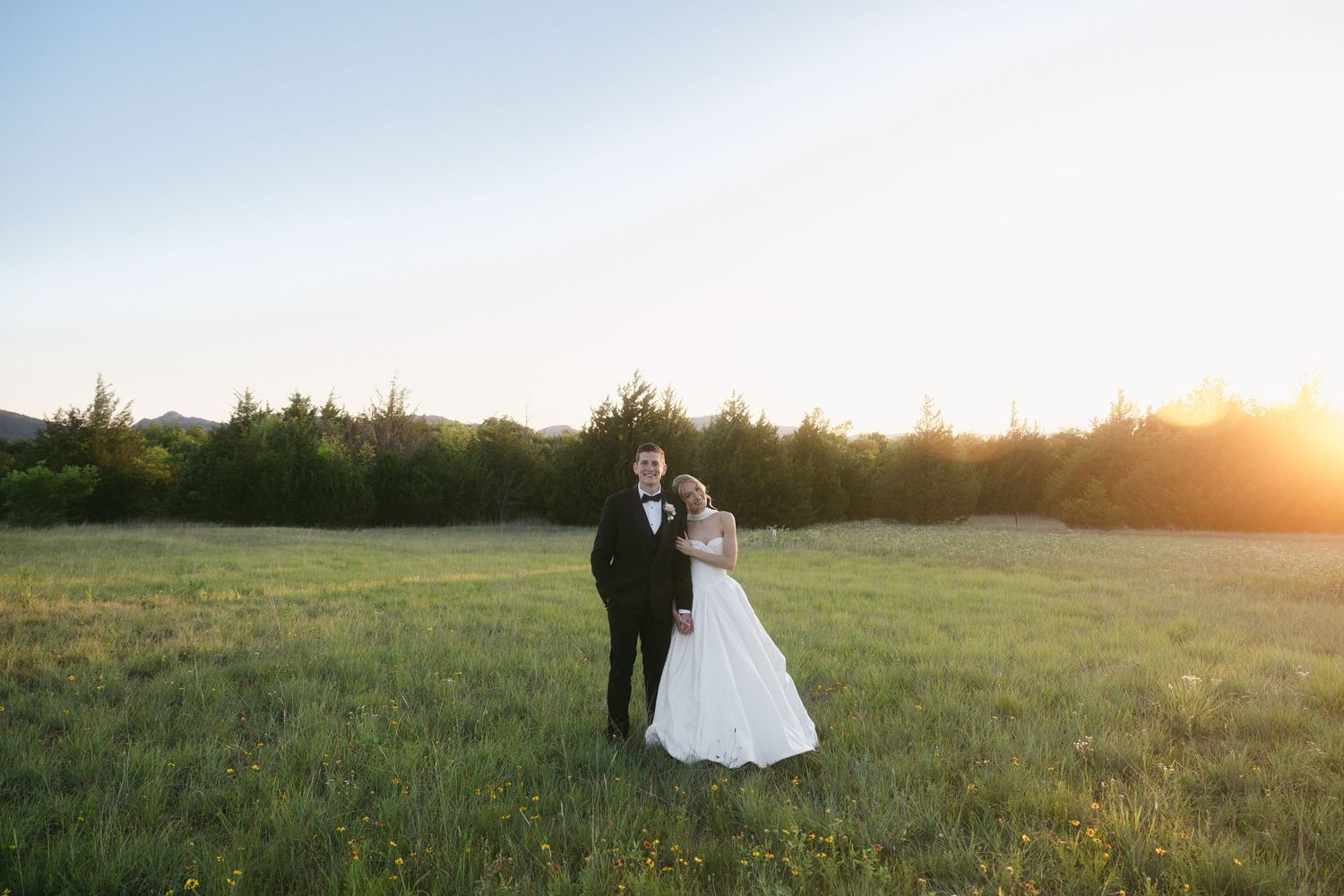 Bride and groom embrace in golden sunset light in a green meadow with trees behind them.