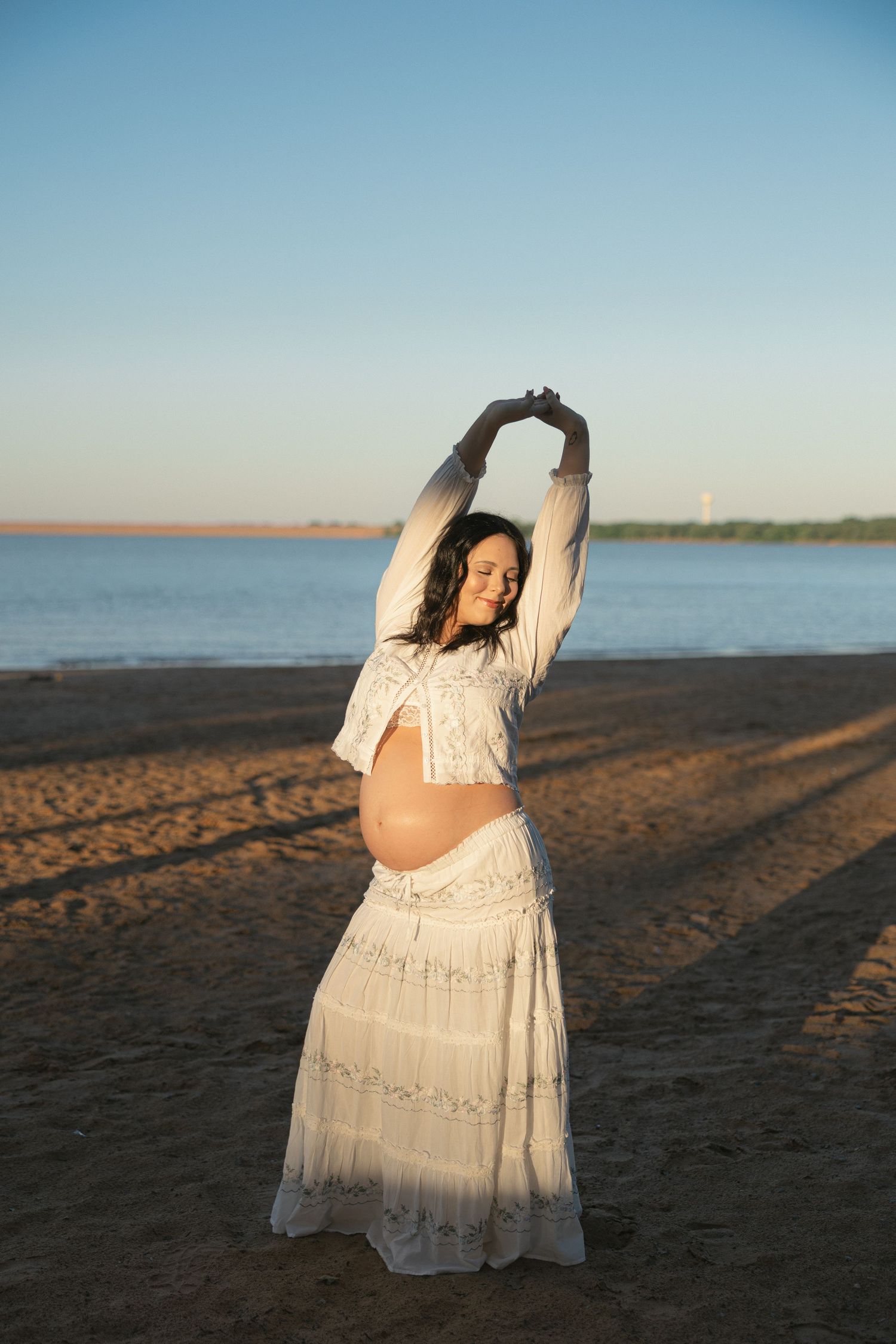 Pregnant woman in white boho dress stretching arms overhead on sandy beach at golden hour sunset.