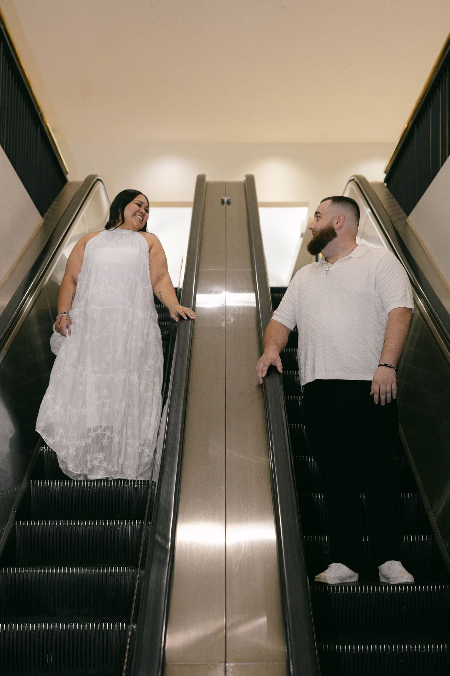 Couple holding hands on escalators, woman in white dress and man in white shirt, smiling at each other.