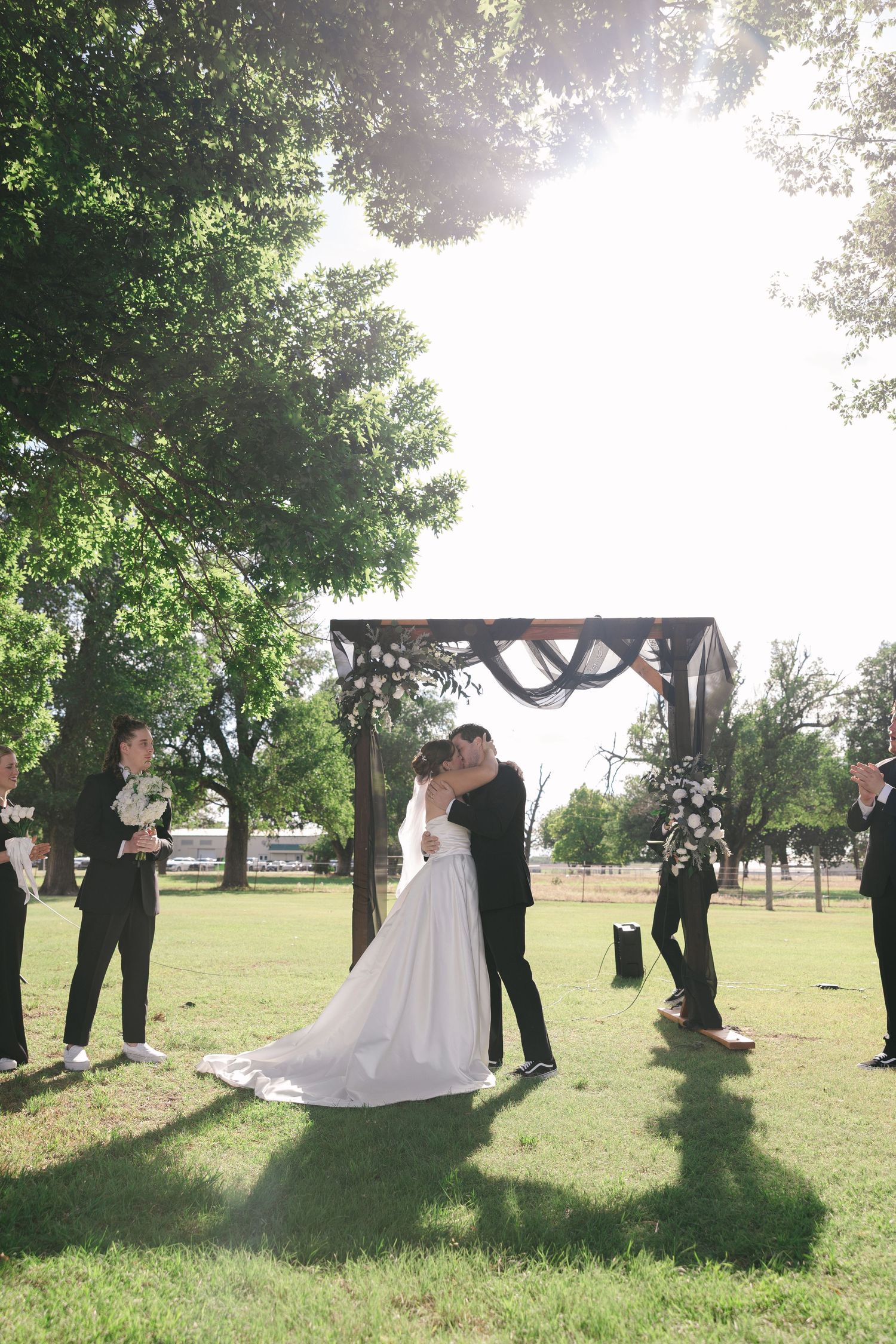 Bride and groom share first kiss under rustic wooden arch outdoors, surrounded by wedding party on sunny green lawn.