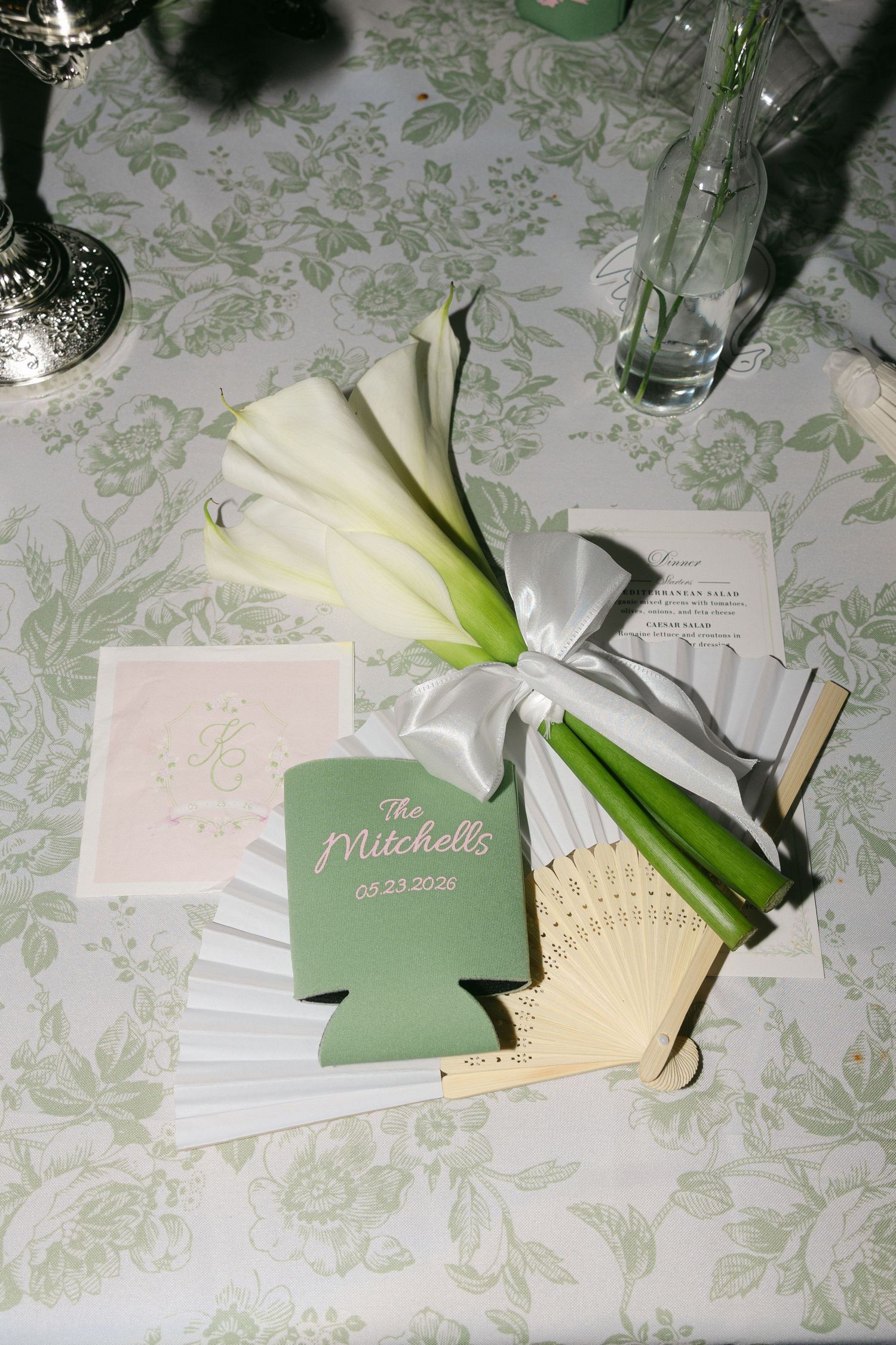 Green and white wedding table setting with calla lilies, a fan, and personalized Mitchell wedding koozies dated 05.23.2025.