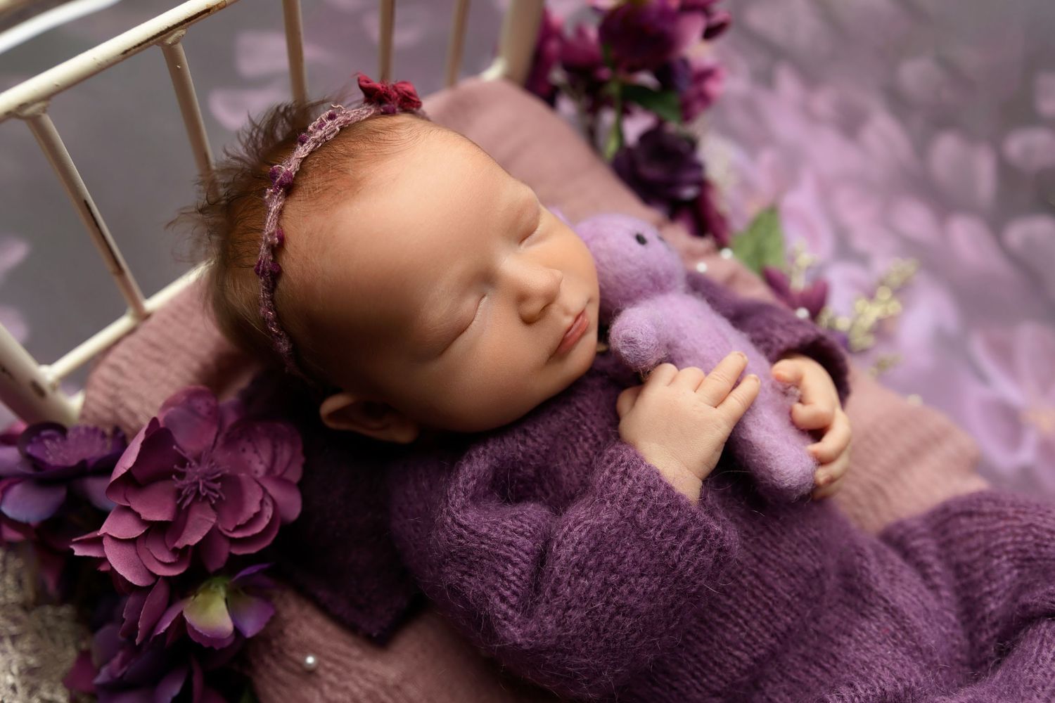 Sleeping newborn baby girl in purple knit wrap with floral headband on a bed with purple flowers.