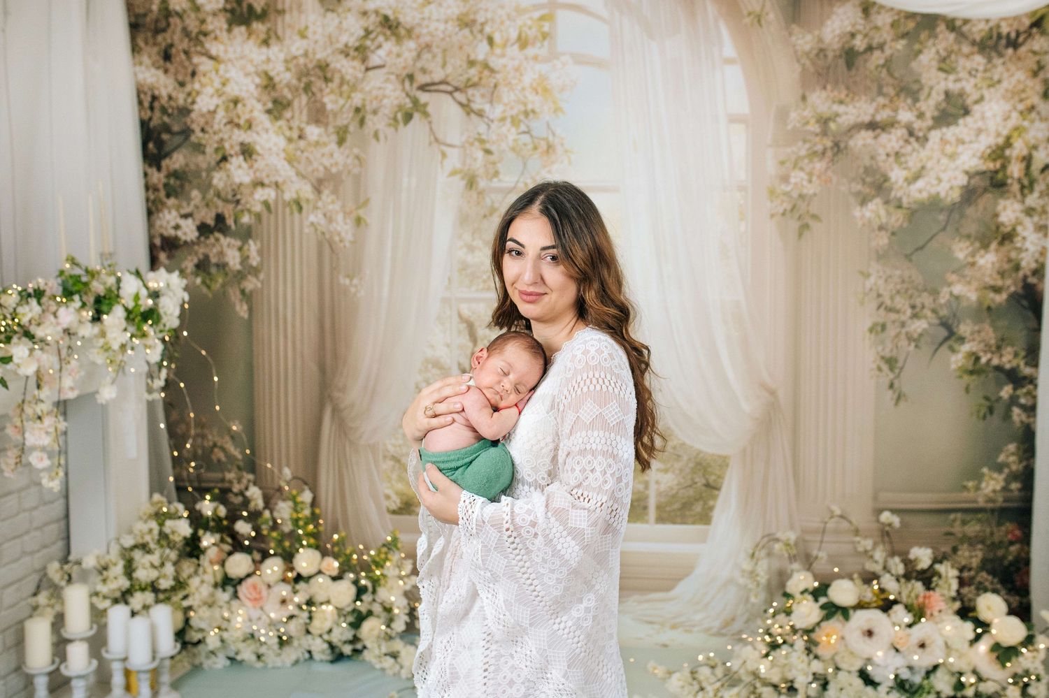 A young woman in a white lace dress holds a sleeping newborn baby in a floral studio setting.