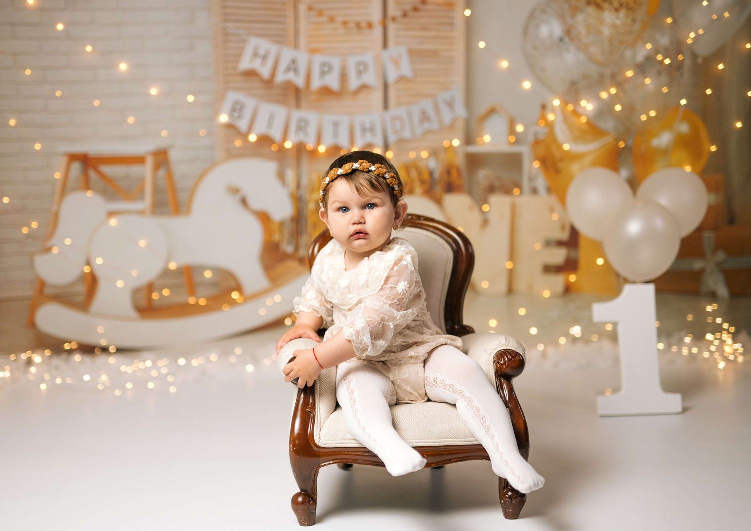 Baby girl in white dress sits in mini chair with birthday banner, balloons, and fairy lights backdrop.