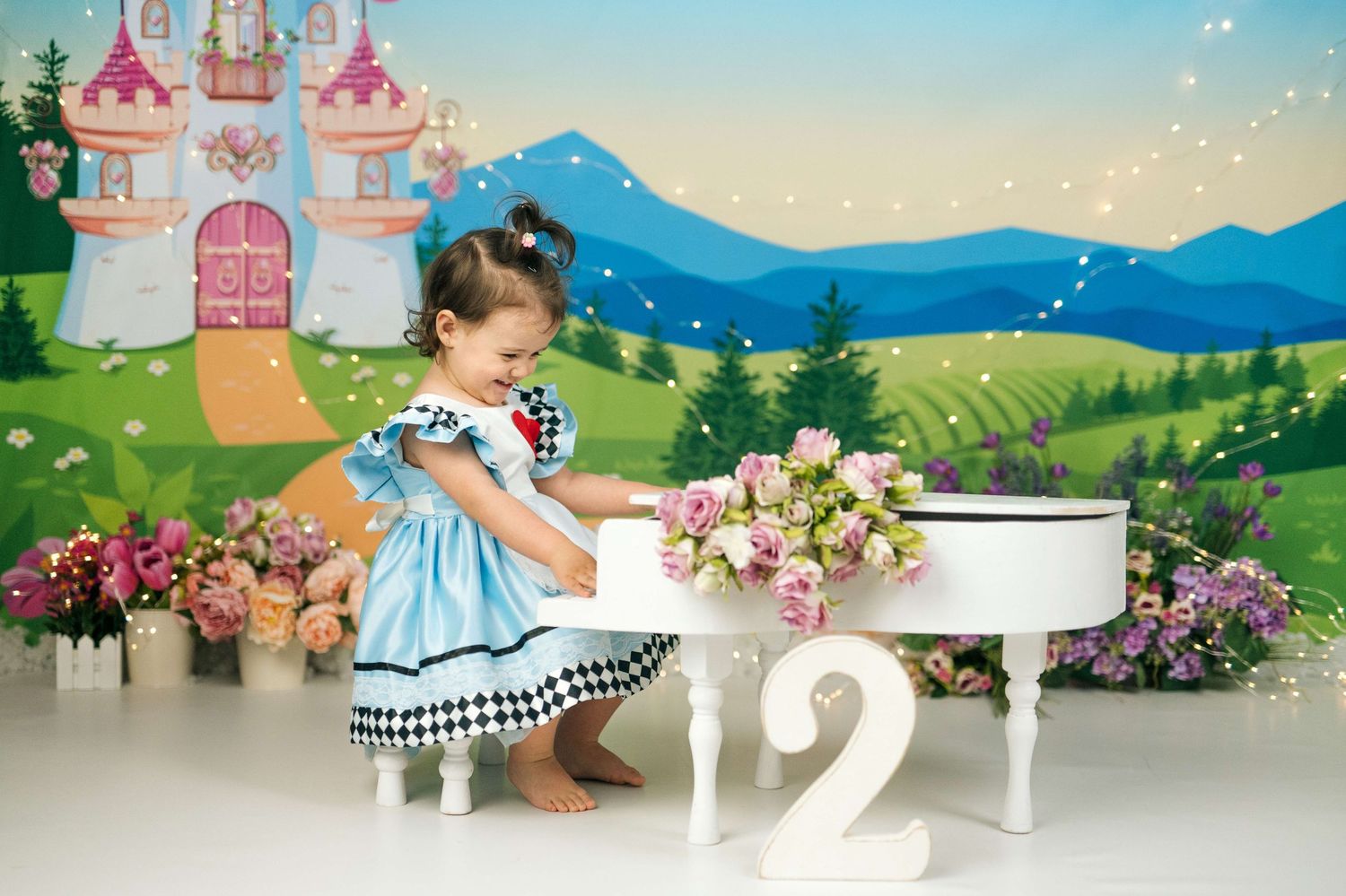 Toddler girl in blue dress poses by white table with flowers, number 2, and castle backdrop for 2nd birthday photo.
