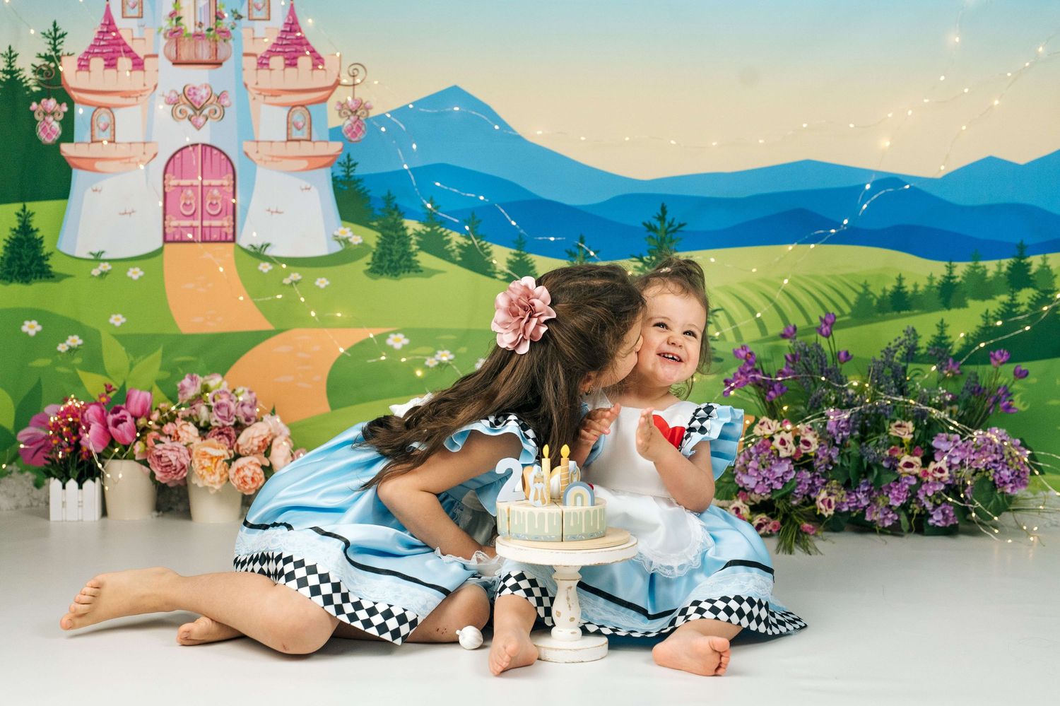 Two young girls in Alice in Wonderland dresses share a sweet kiss near a birthday cake with a fairy tale castle backdrop.