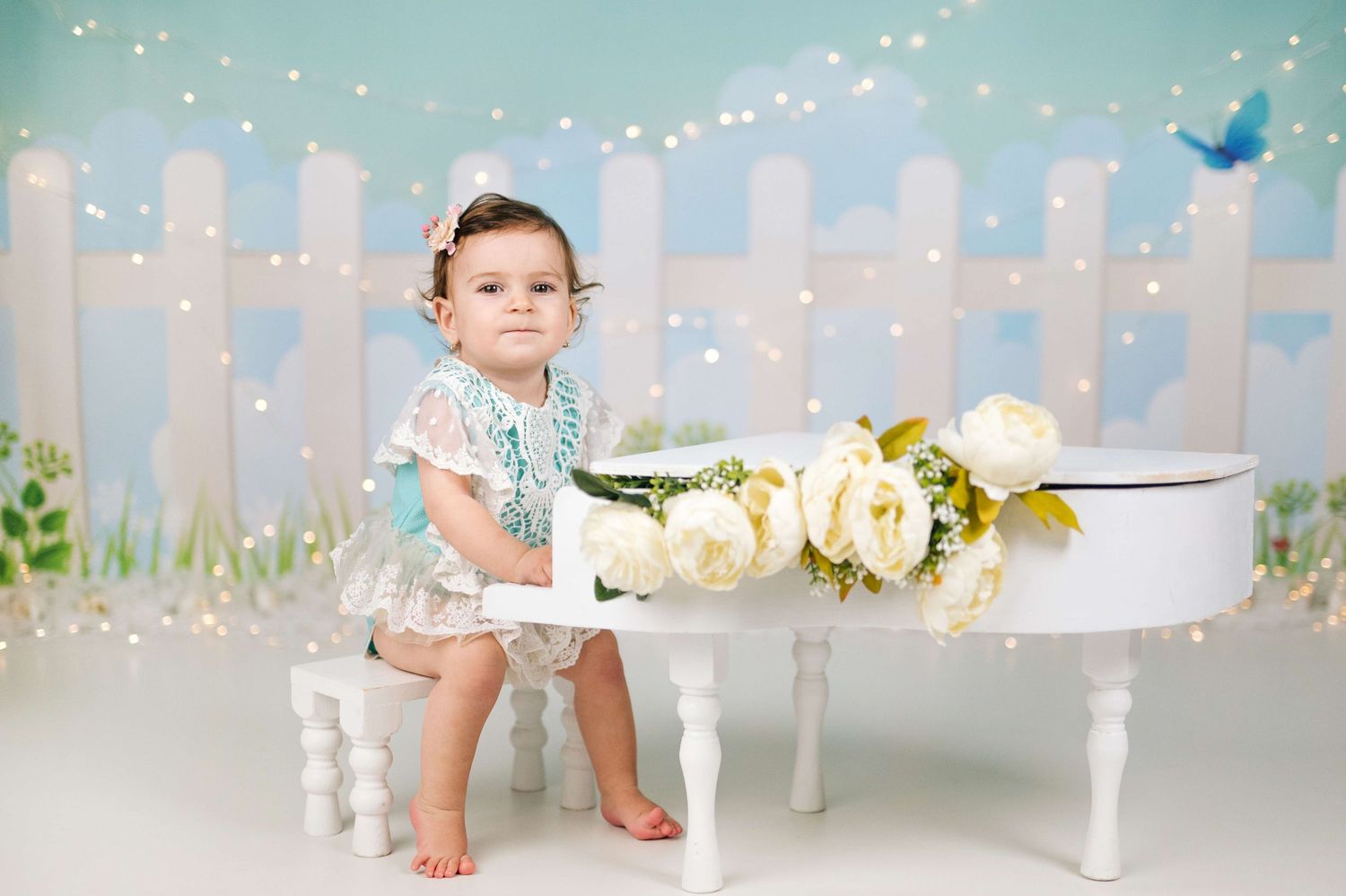 Adorable toddler girl in lace dress sits beside white mini piano with flowers for first birthday photos.