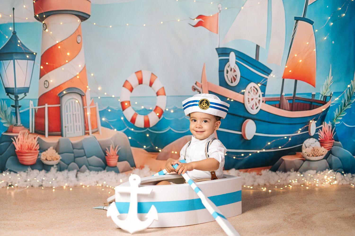 Baby boy in sailor outfit sits in toy boat with nautical backdrop featuring lighthouse and ship.