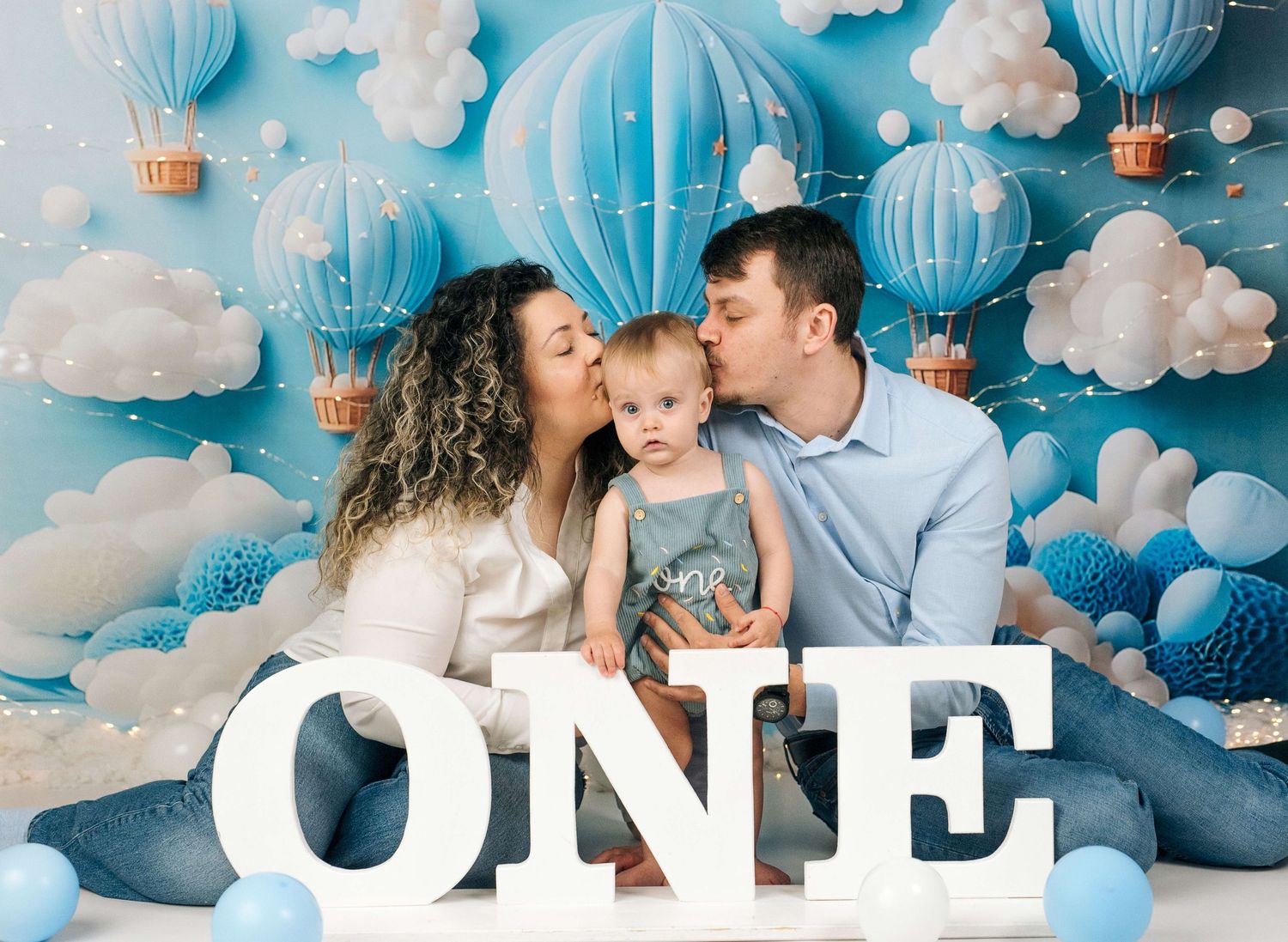 Family celebrating baby's first birthday with blue hot air balloon backdrop and large ONE letters.