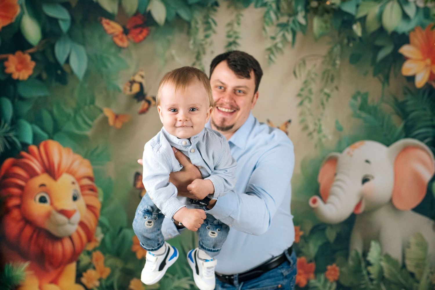 Father holding smiling baby boy in front of colorful jungle backdrop with lion and elephant.
