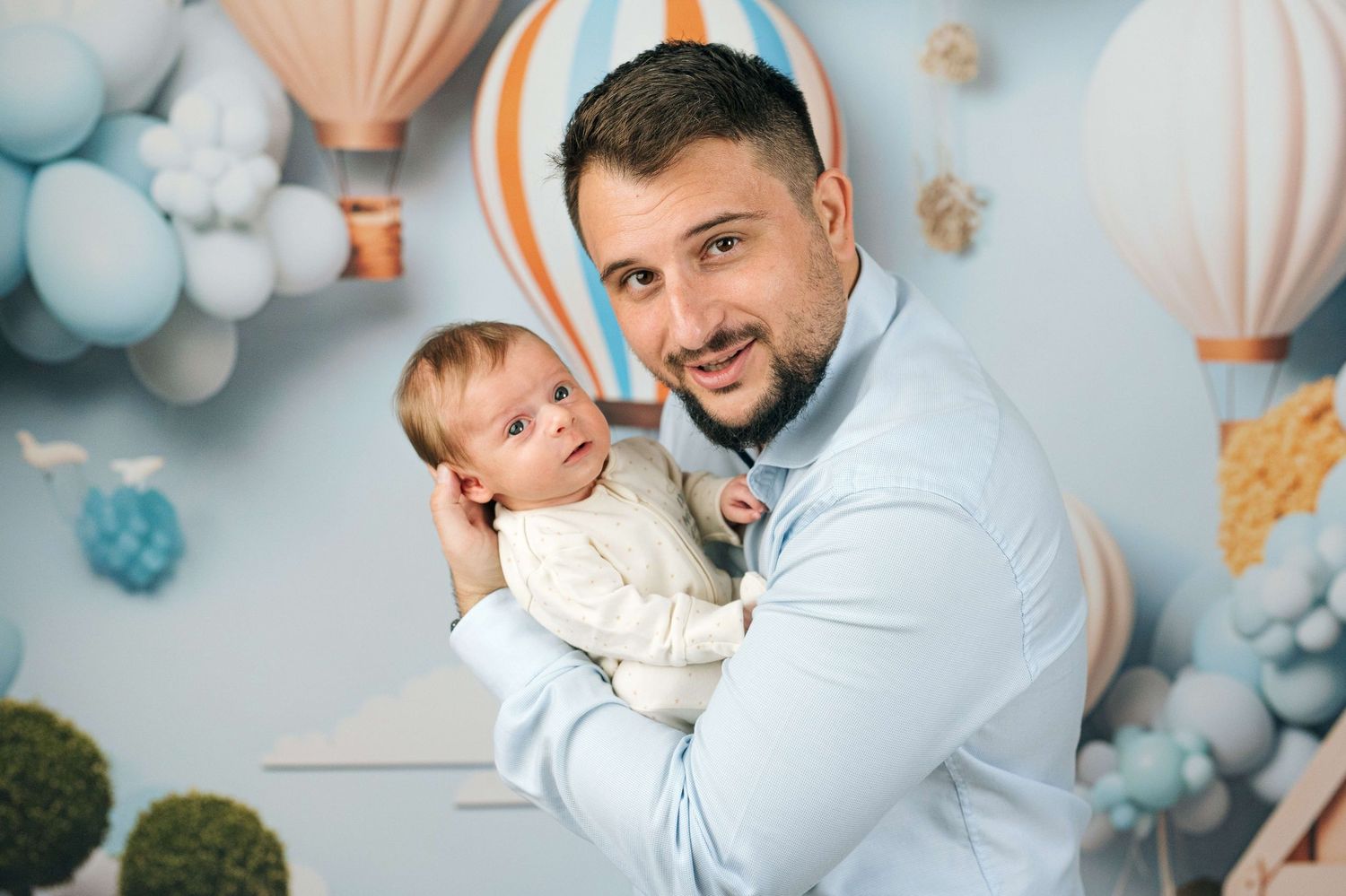 A smiling man holds a baby in front of a whimsical hot air balloon backdrop with blue tones.