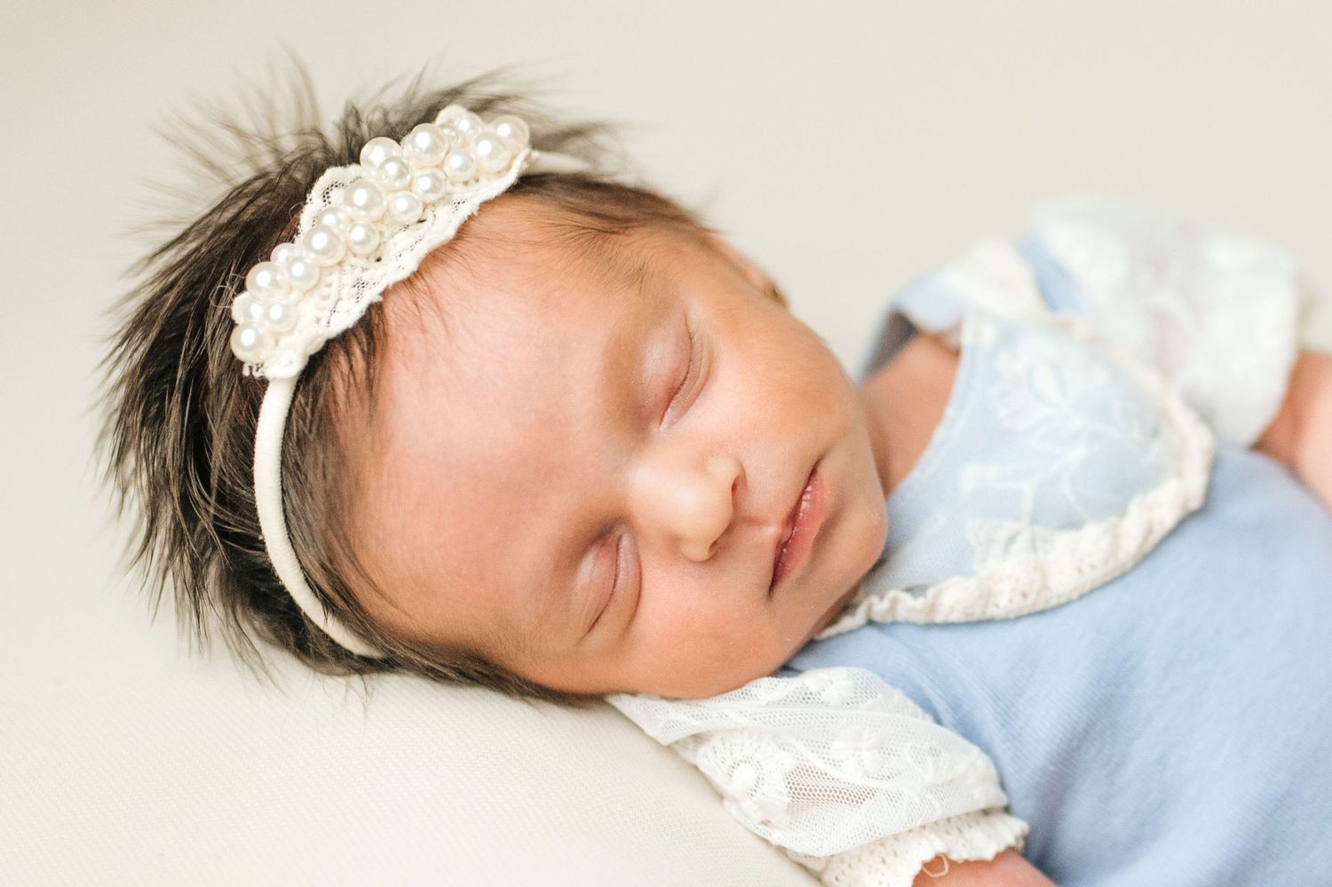 Sleeping newborn baby girl wearing a white pearl headband and blue lace outfit on a soft white background.