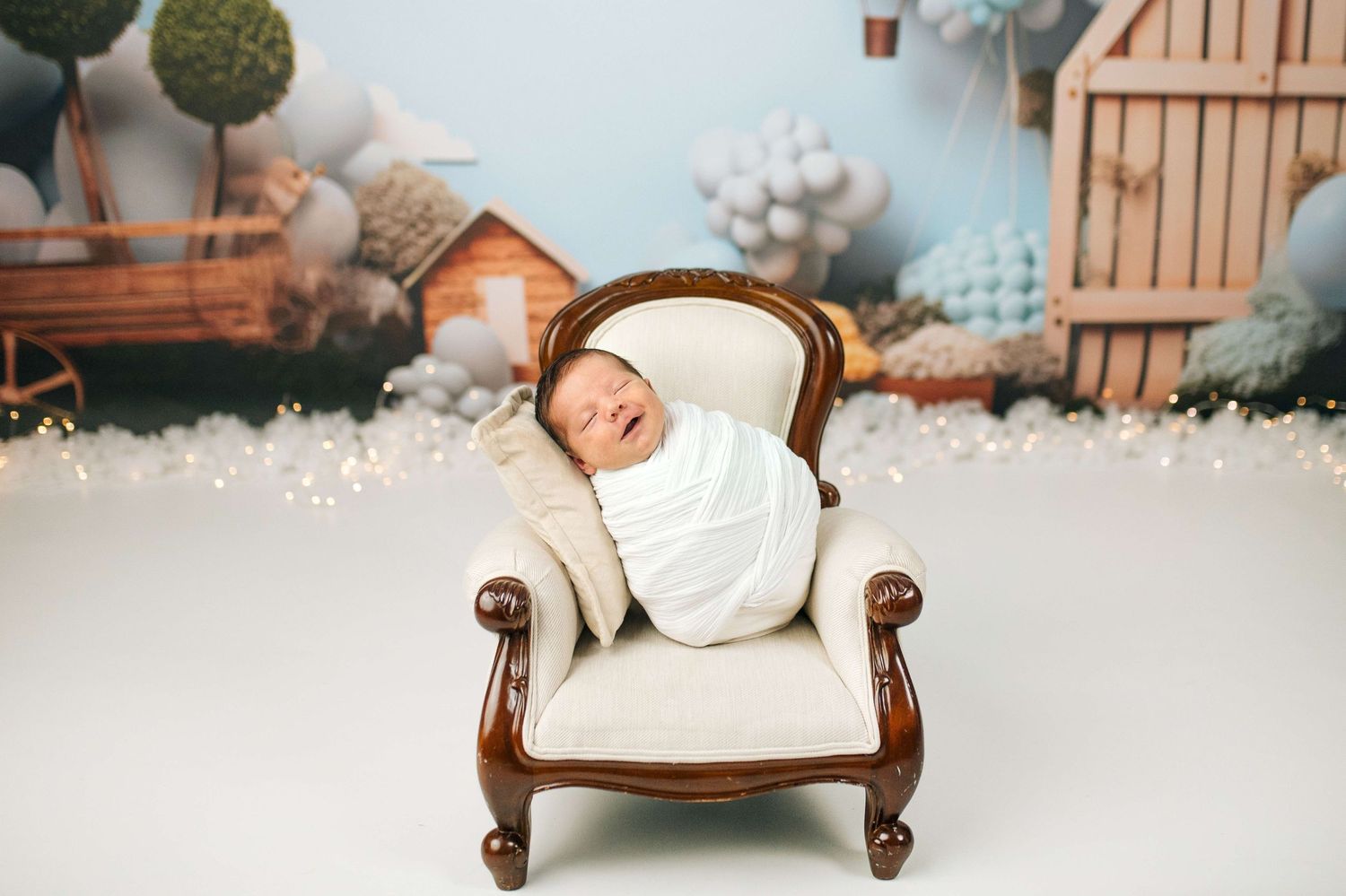 Sleeping newborn baby swaddled in white on a miniature armchair in a charming studio photography setup.