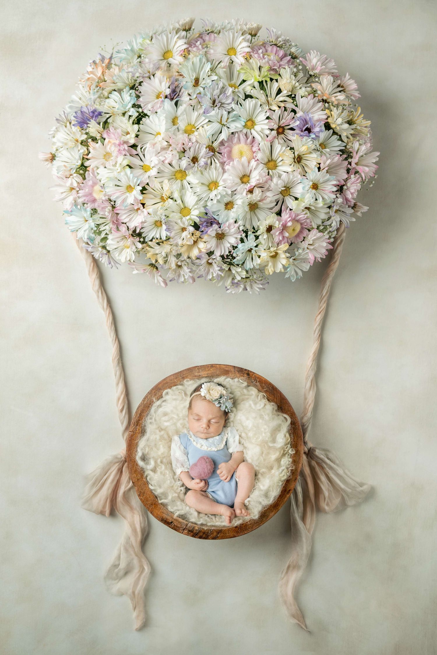 Newborn baby sleeping in a wooden bowl suspended from a floral hot air balloon made of daisies in pastel colors.