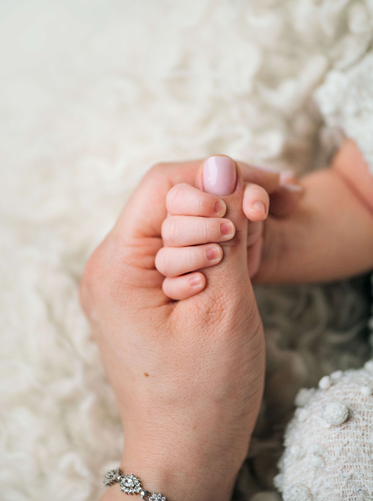 A mother gently holds her newborn baby's tiny hand, wearing a delicate bracelet, on a soft white blanket.
