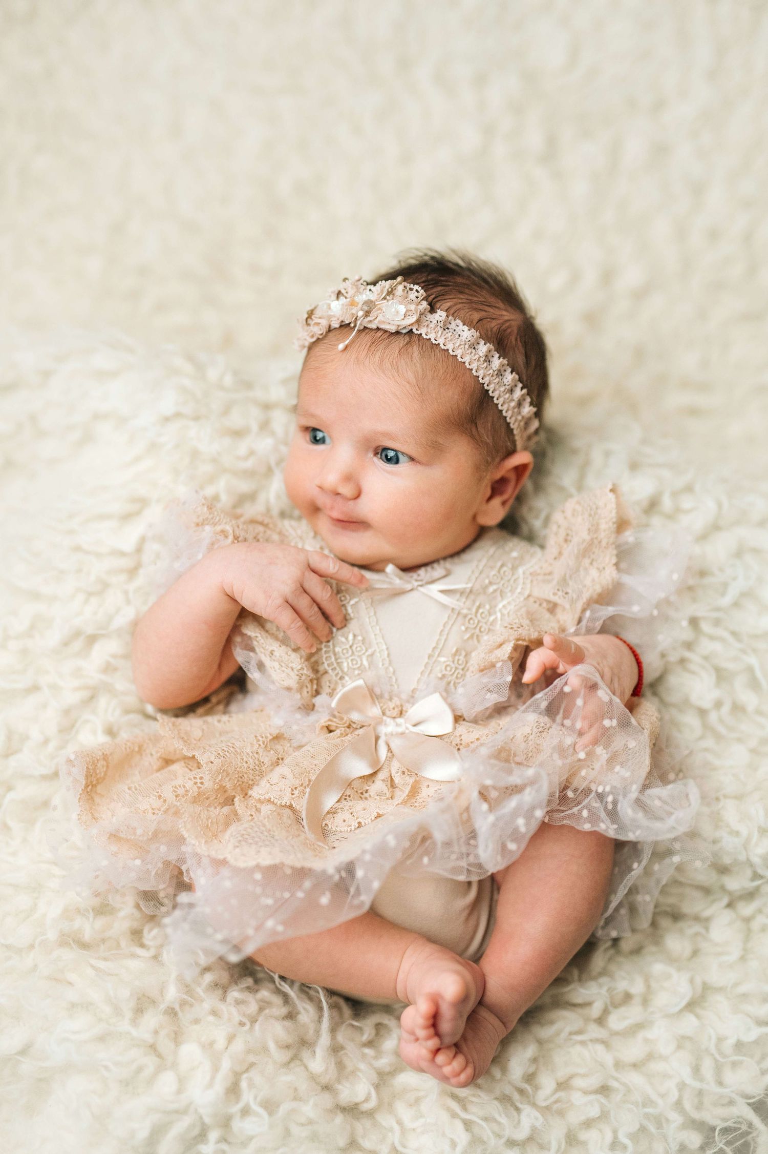 Adorable baby girl in a cream lace dress and floral headband sitting on a fluffy white rug.