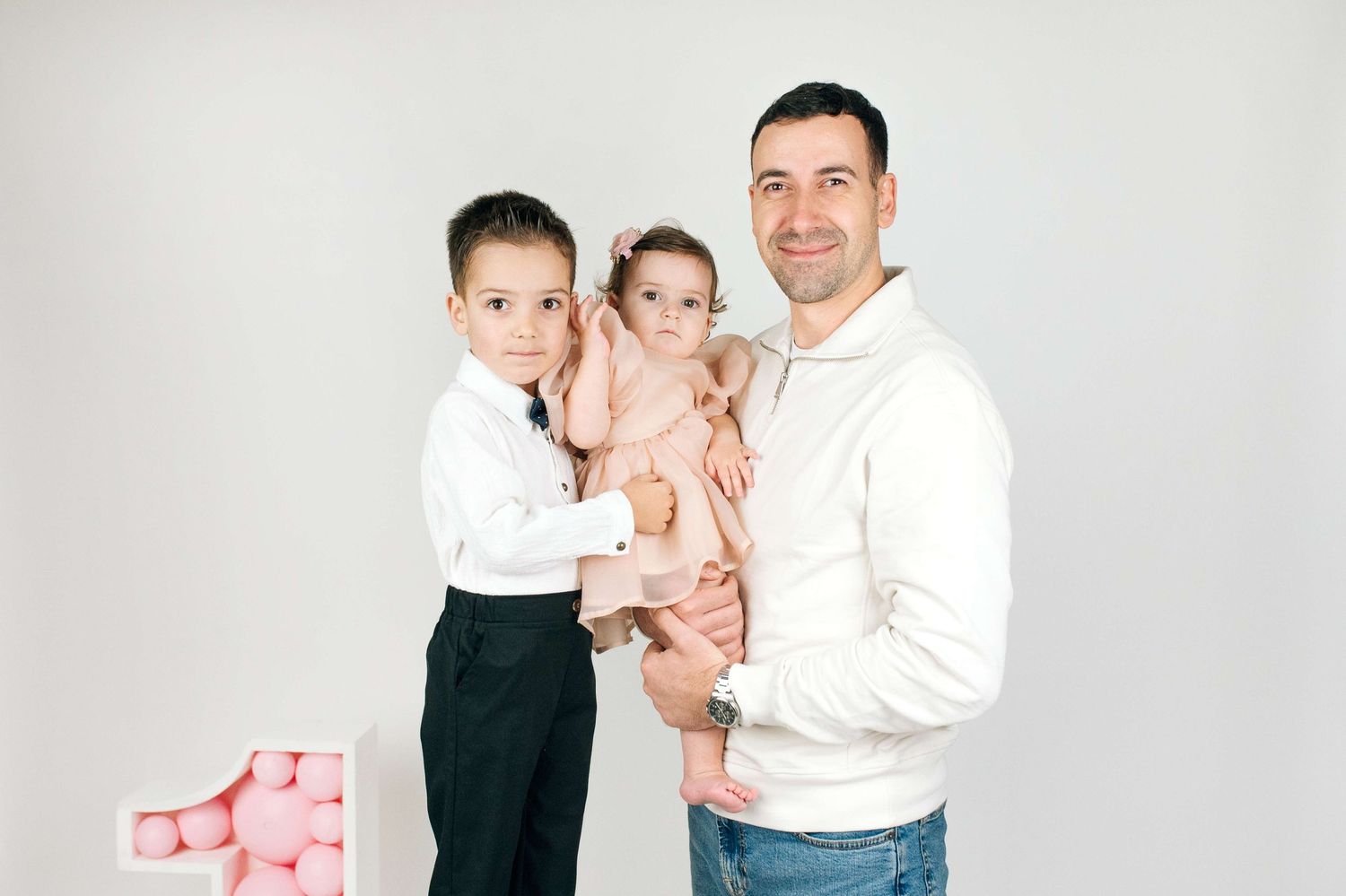 A man in a white hoodie holds a baby girl while a young boy in a white shirt and black pants stands beside them.