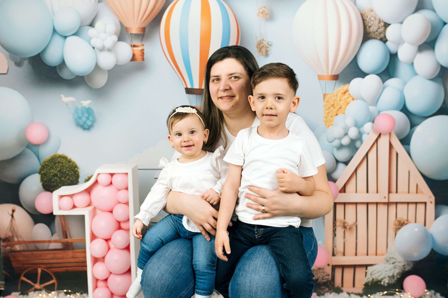 A mother poses with her two young children in front of a colorful balloon and hot air balloon themed backdrop.