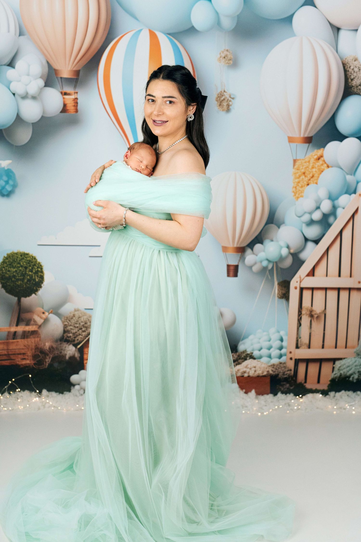 A woman in a mint green gown holds a newborn baby, surrounded by hot air balloon decorations in soft blue tones.