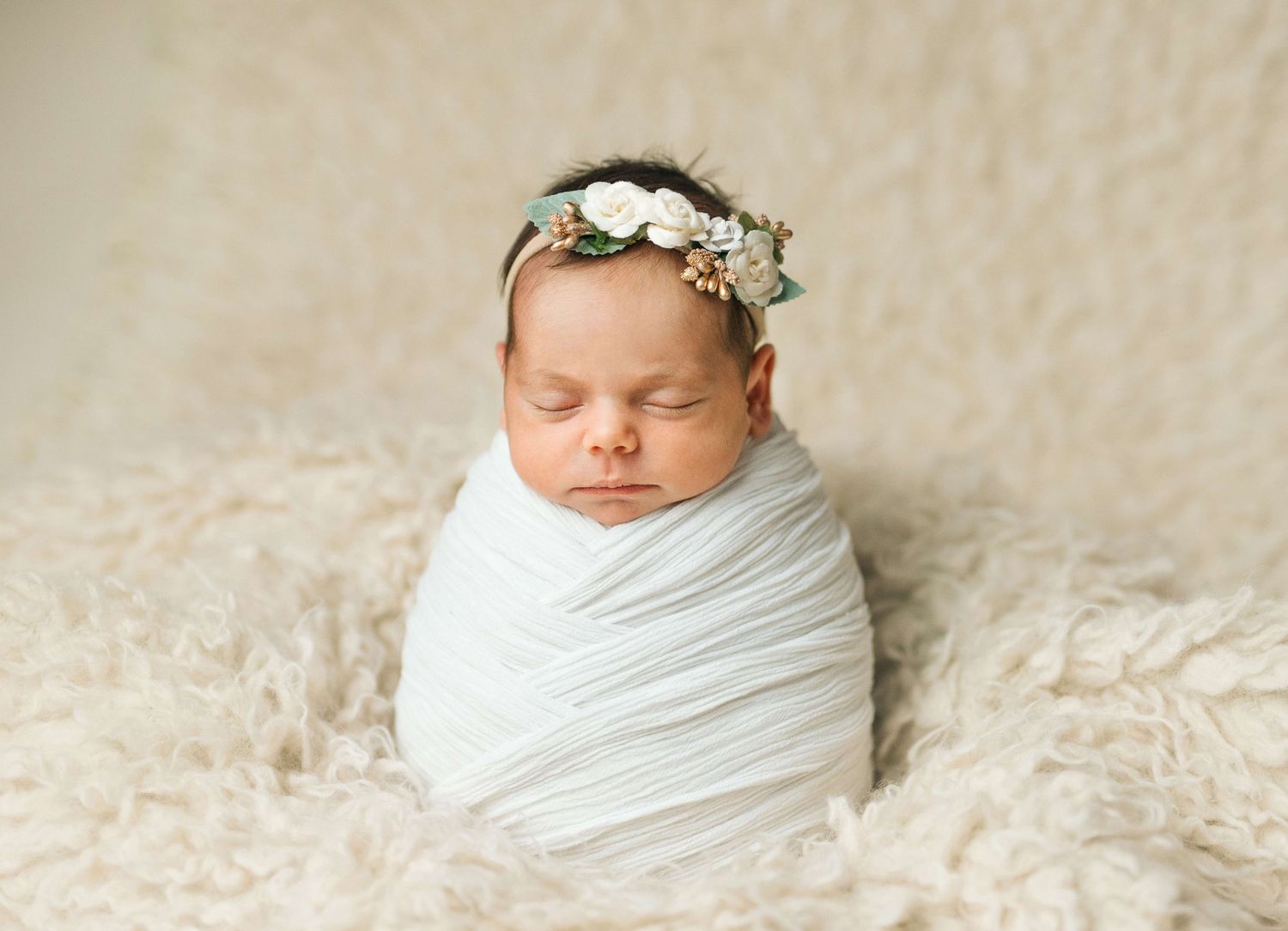 Sleeping newborn baby swaddled in white wrap with floral headband on fluffy cream backdrop.